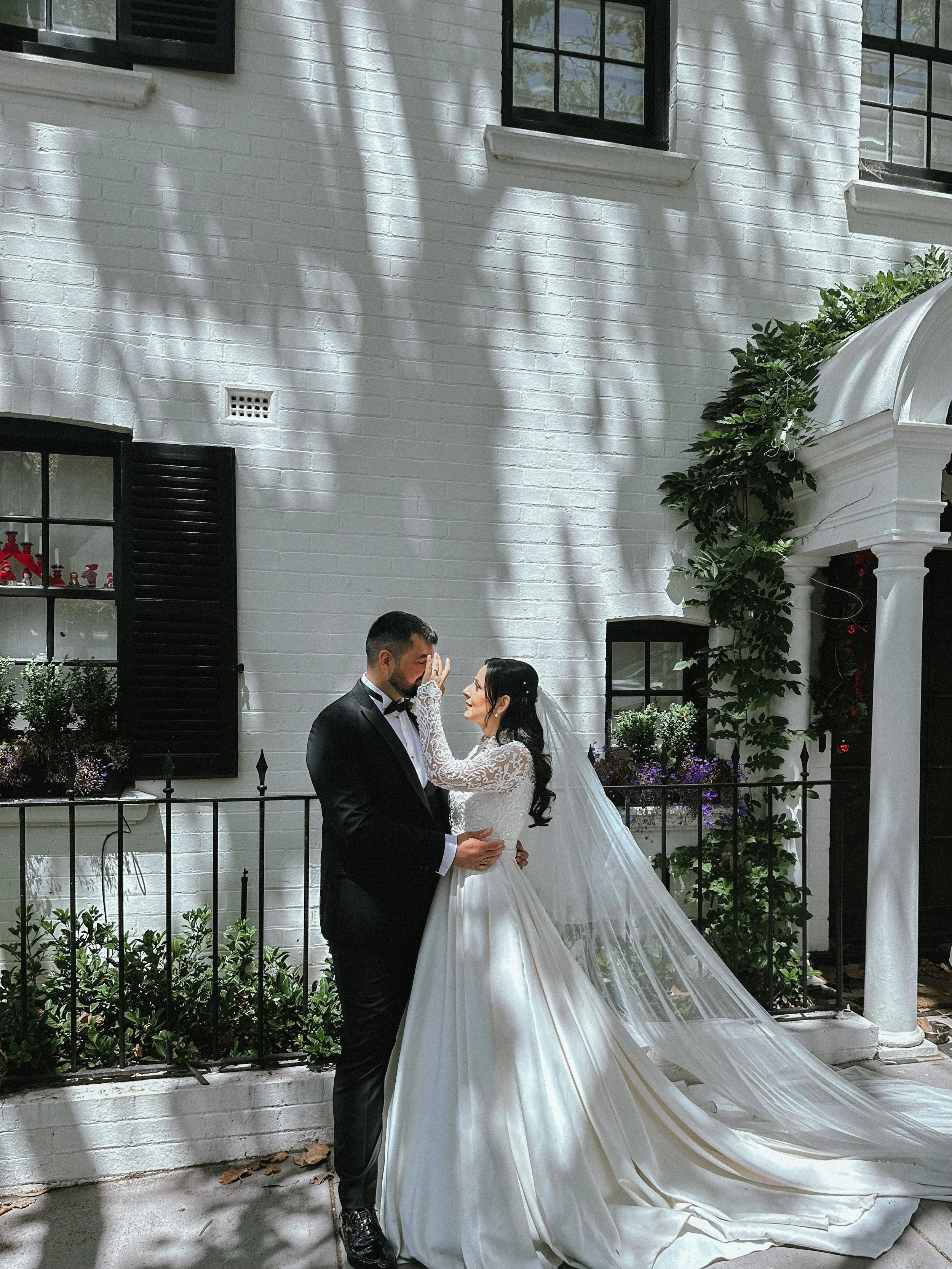 A bride and groom sharing a romantic moment outdoors against a white brick wall with shadows of tree branches. The bride is wearing a white wedding gown with a long veil, and the groom is in a black tuxedo.