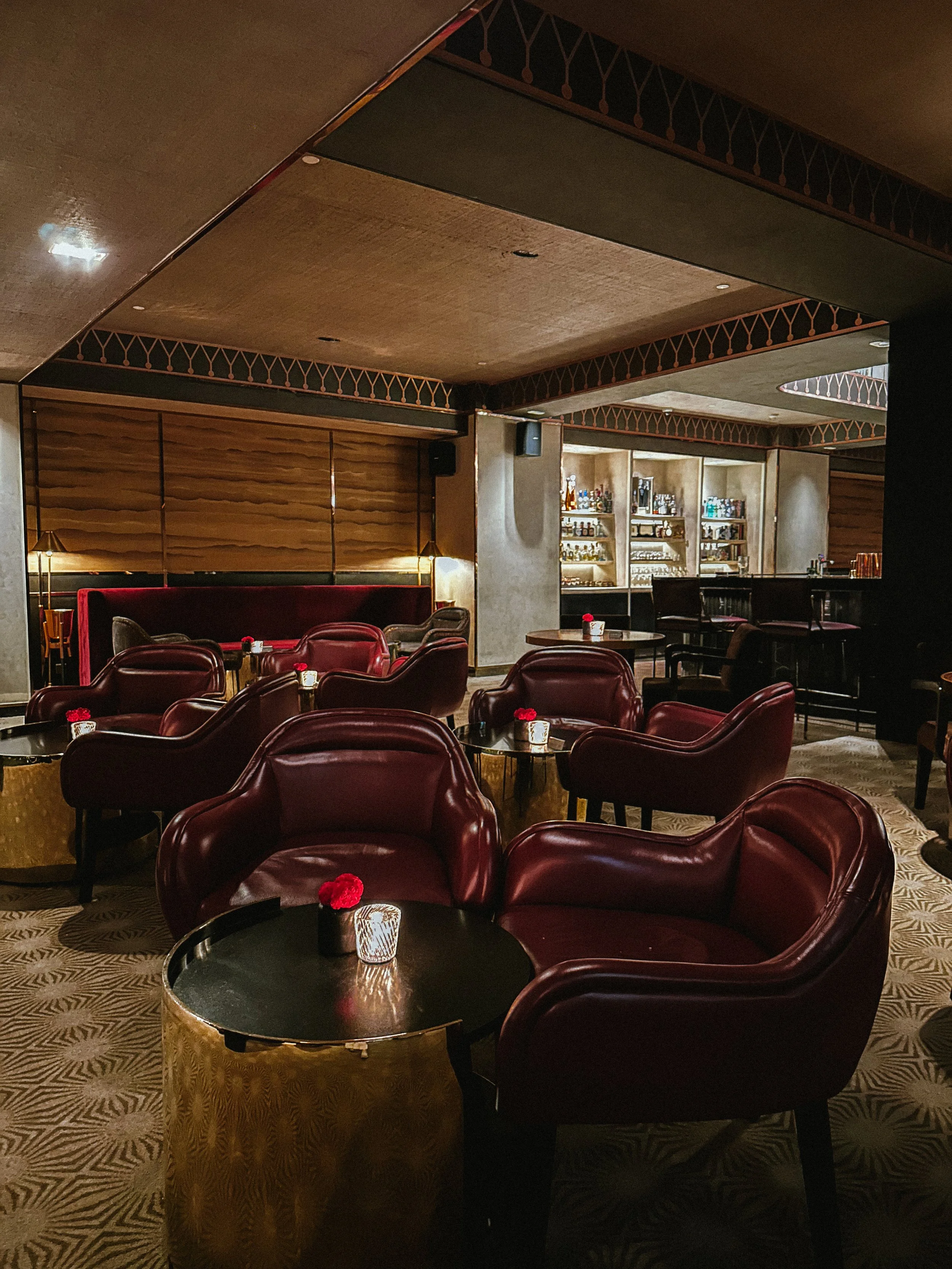 Interior of a lounge bar with red leather armchairs, small tables decorated with red flowers and candles, and a bar in the background with liquor bottles on shelves.