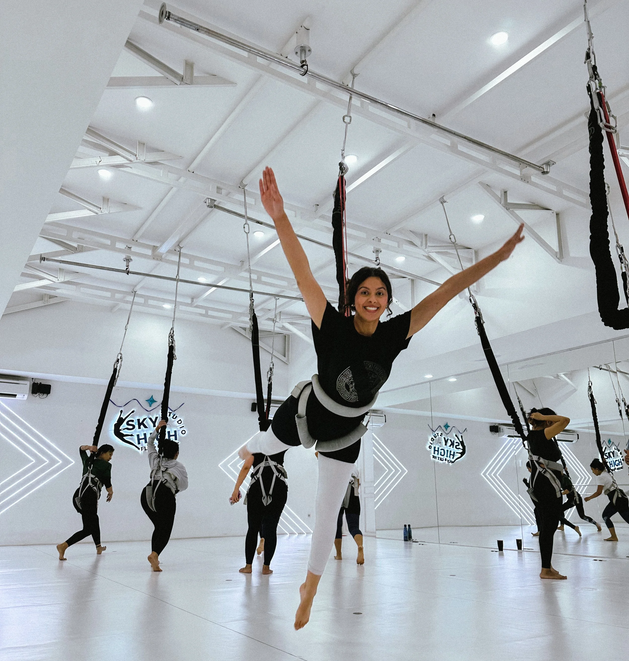 A woman smiling and posing while hanging from aerial silks in a fitness studio, with other participants in the background practicing aerial exercises.