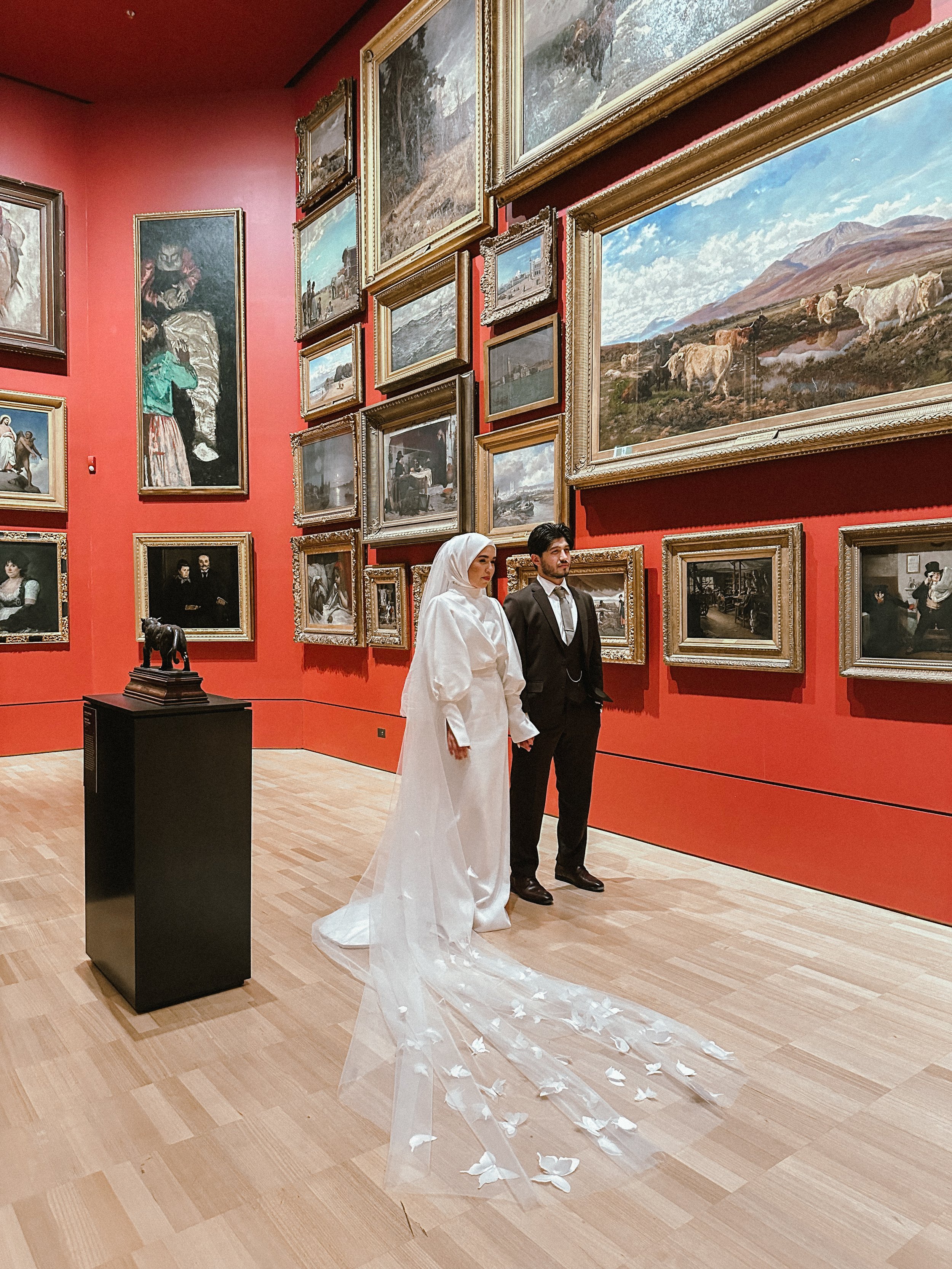 A bride and groom holding hands in an art gallery with framed paintings on red walls, and a small sculpture on a black pedestal, with the bride wearing a white wedding dress and veil, and the groom in a black suit.