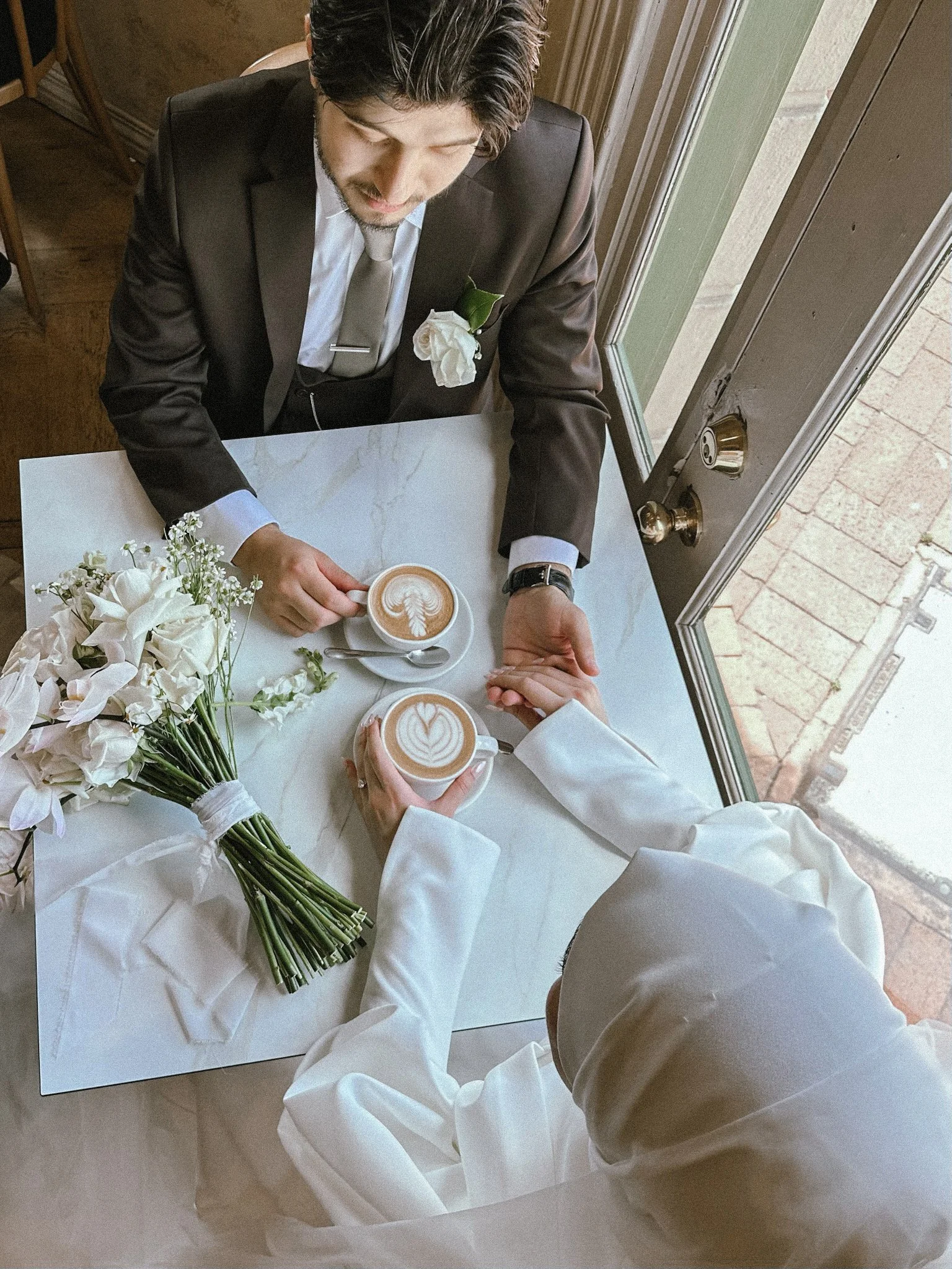 A man in a black suit with a white boutonniere sitting at a white marble-topped table, holding a coffee cup with latte art, and a woman in a white blazer sitting across with her hand on his, also holding a latte with latte art. A bouquet of white flo