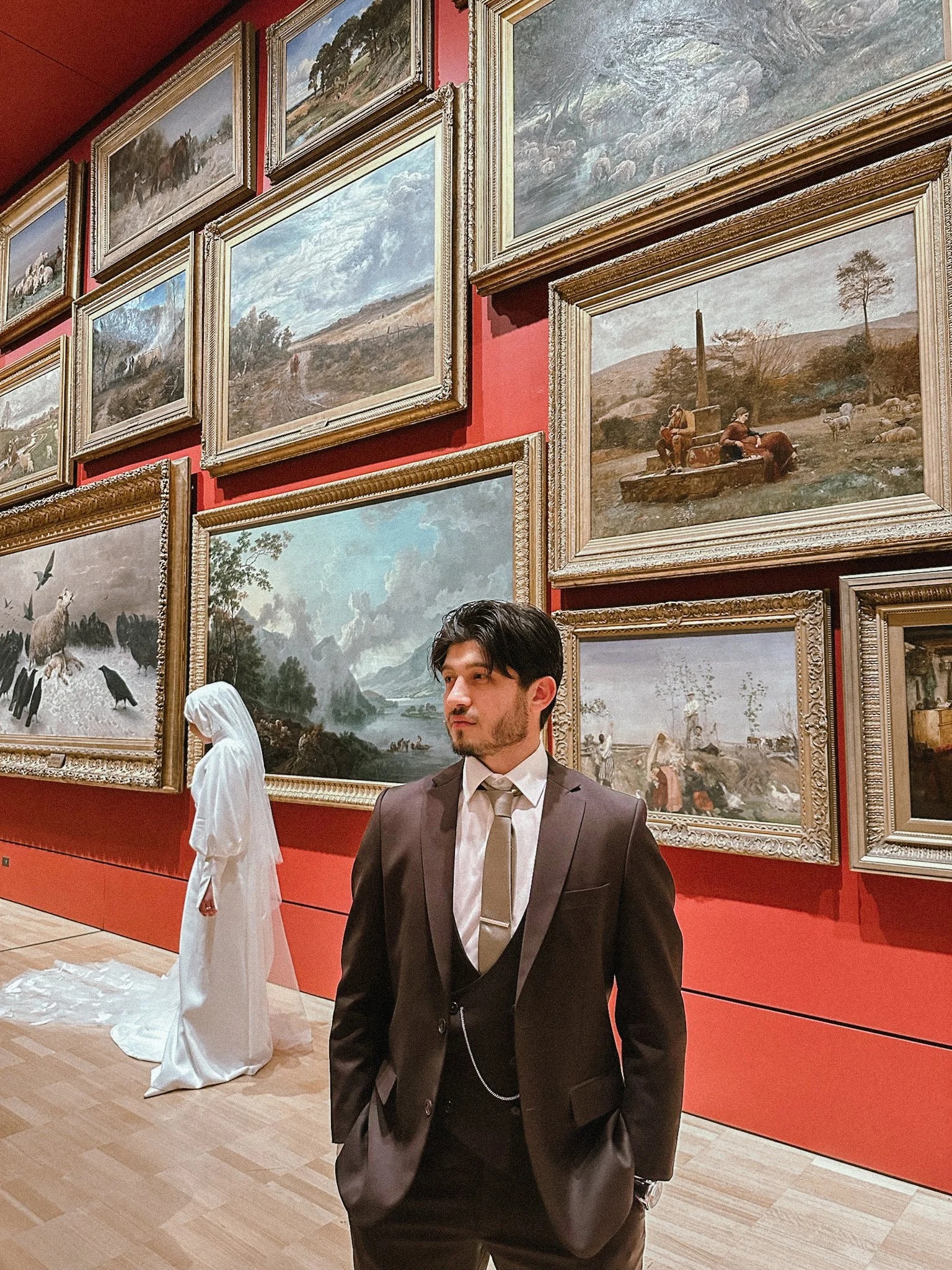 A man in a dark suit and tie stands in front of a wall with many framed landscape paintings at an art gallery or museum, and a figure dressed in white is visible in the background near the paintings.