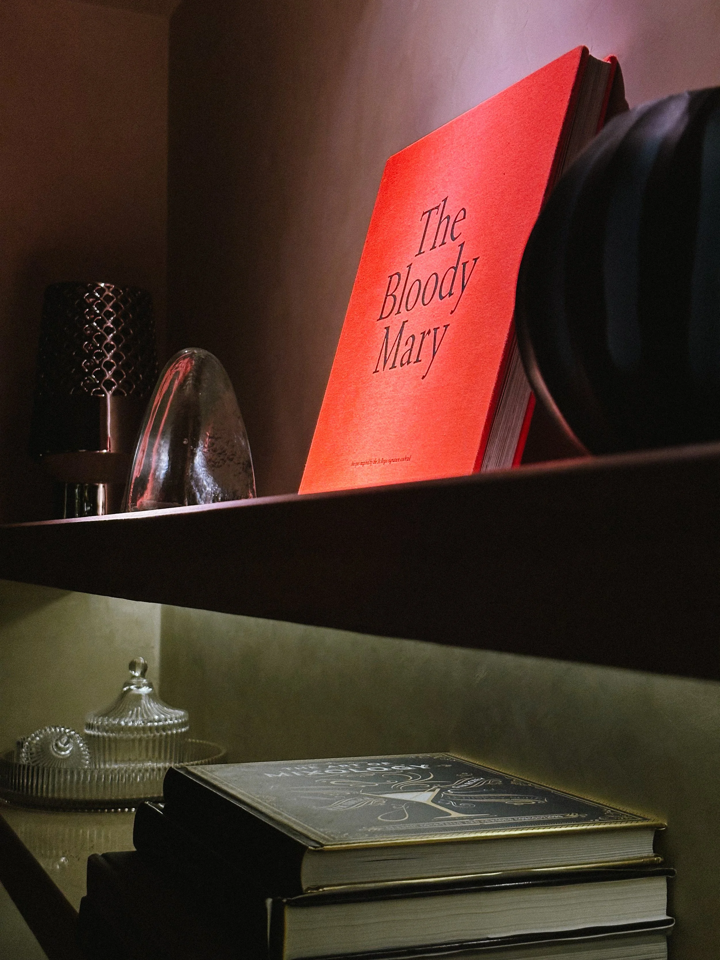 A bookshelf with a red book titled 'The Bloody Mary', two stacked books underneath, and a decorative glass container with a lid, along with metallic objects on the shelf.