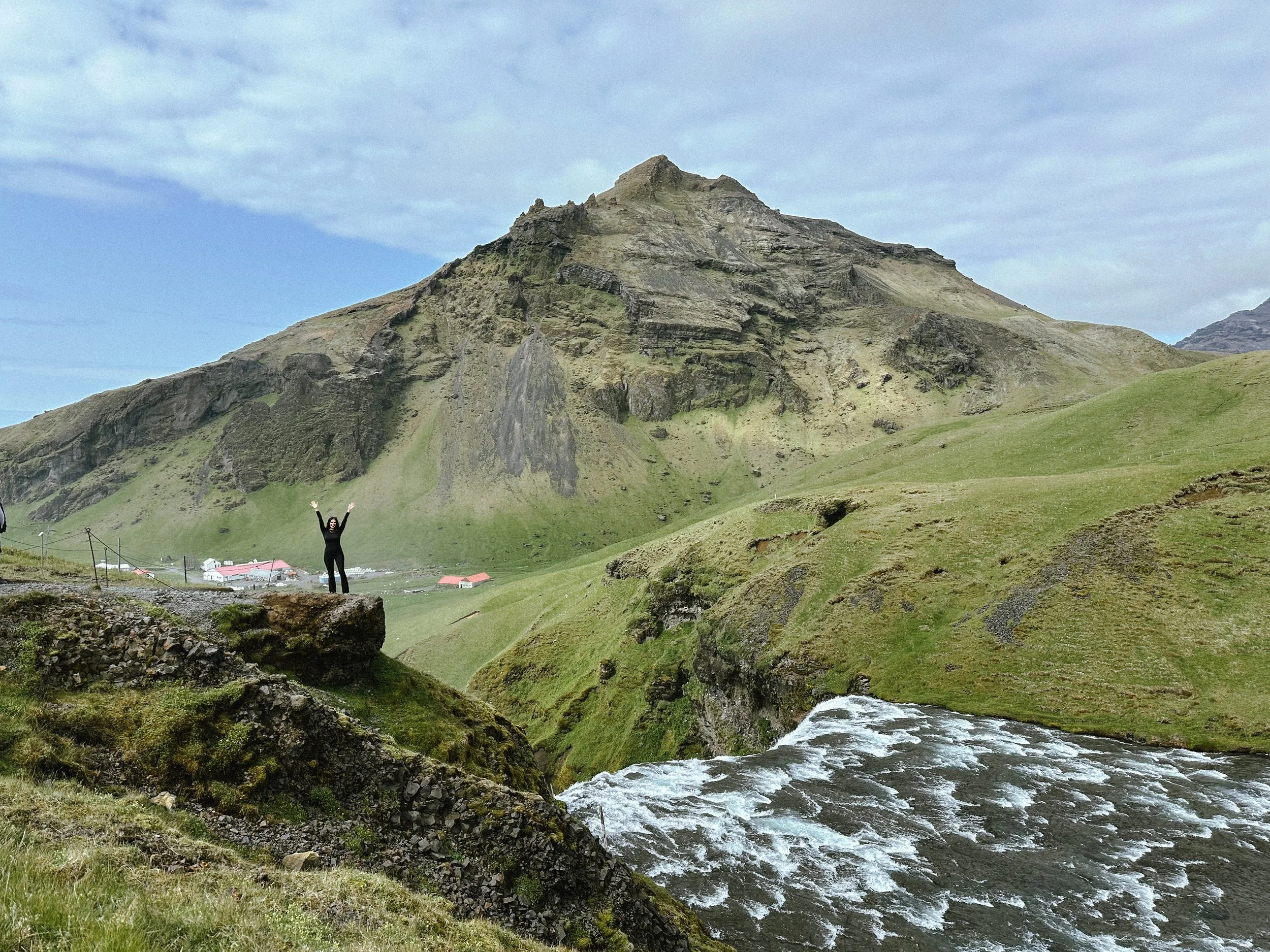 A person standing on a rock with arms raised, overlooking a grassy valley with a/stream, and a large mountain in the background under a partly cloudy sky.