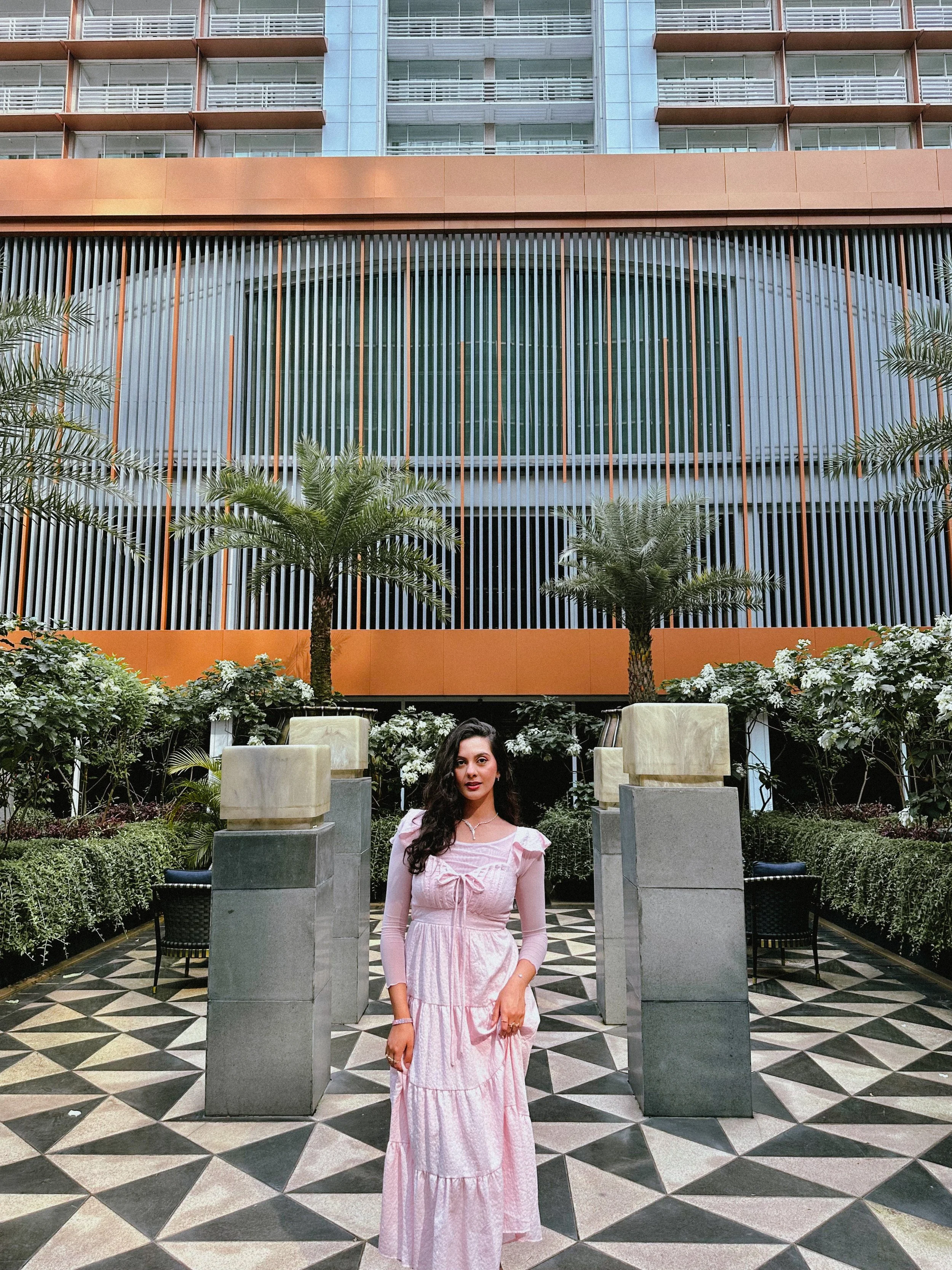 A woman in a pink dress standing in an outdoor courtyard with patterned tile ground, surrounded by greenery, palm trees, and modern building architecture with large windows and vertical metal slats in the background.