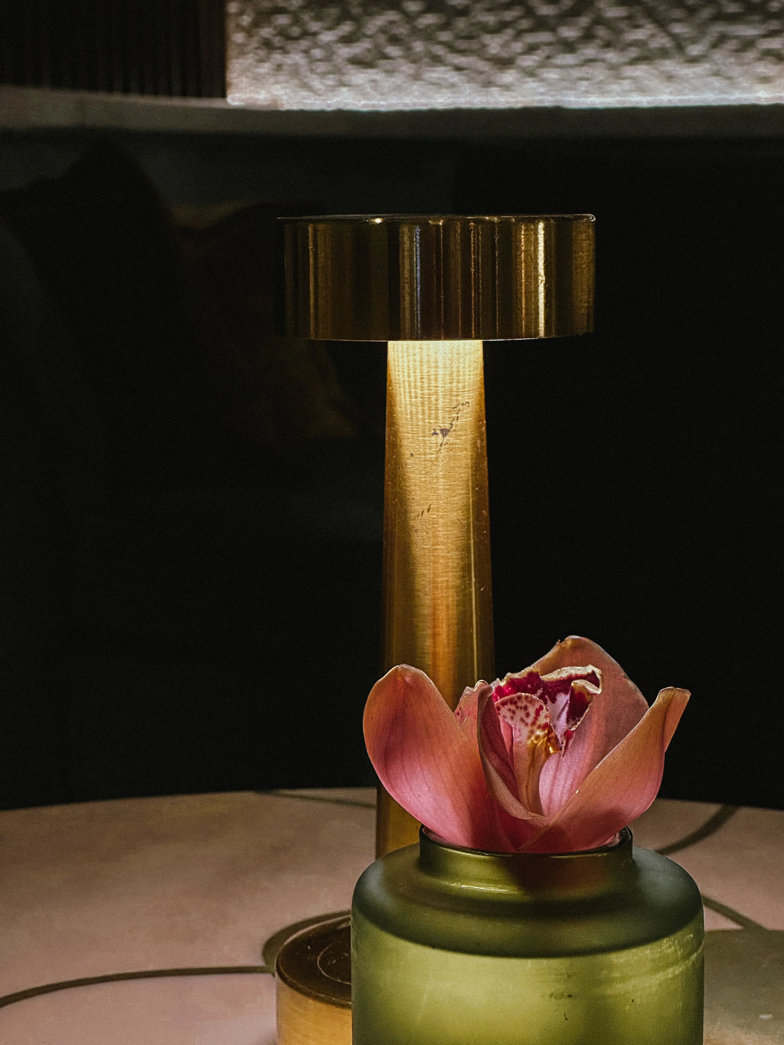 A decorative lamp with a metallic cylindrical shade and a tall metal stem, placed on a table with a pink flower in a green vase in front of it.