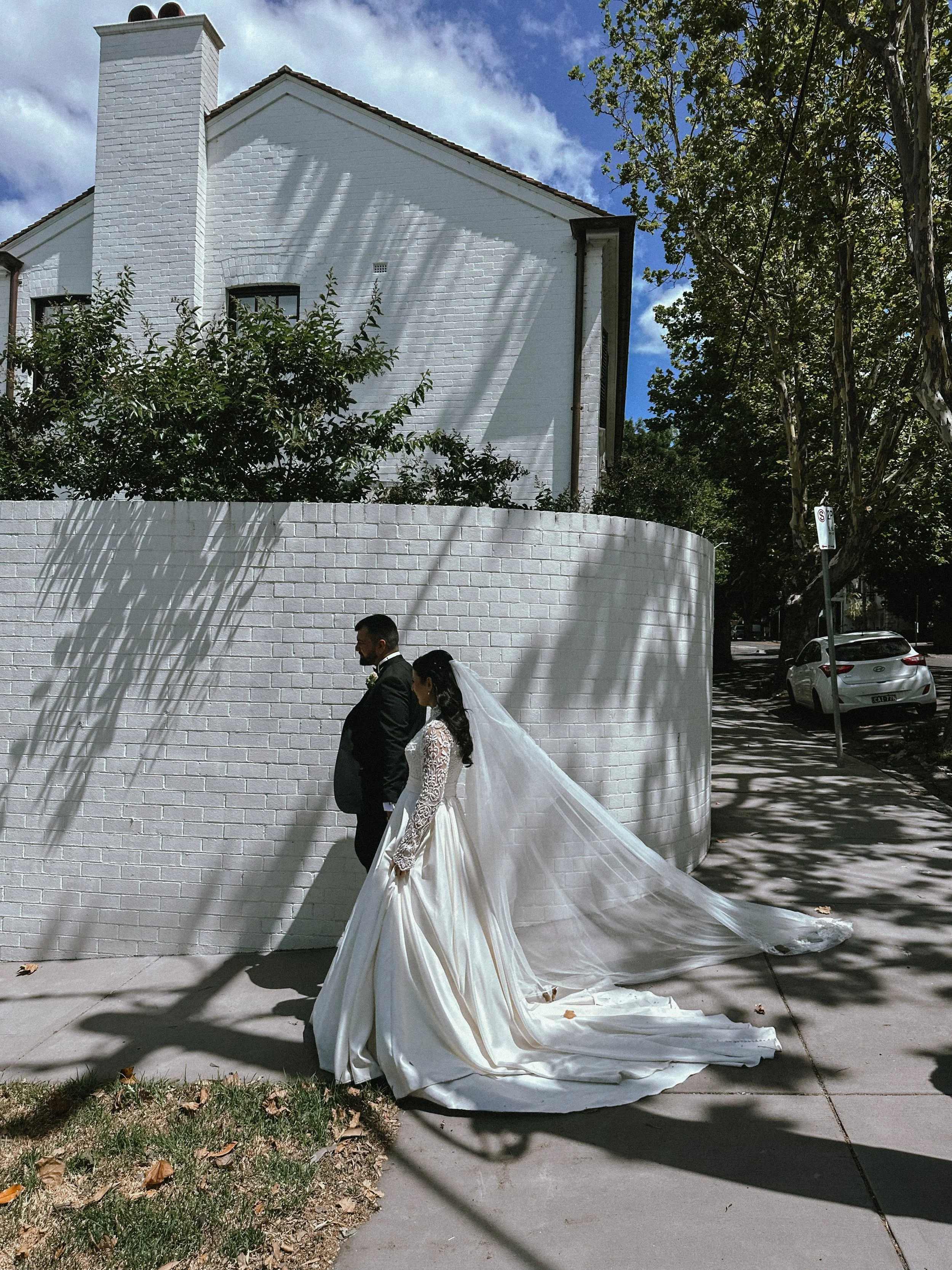 A bride and groom walking on the sidewalk next to a white brick wall, with shadows of tree branches overhead, during daytime.