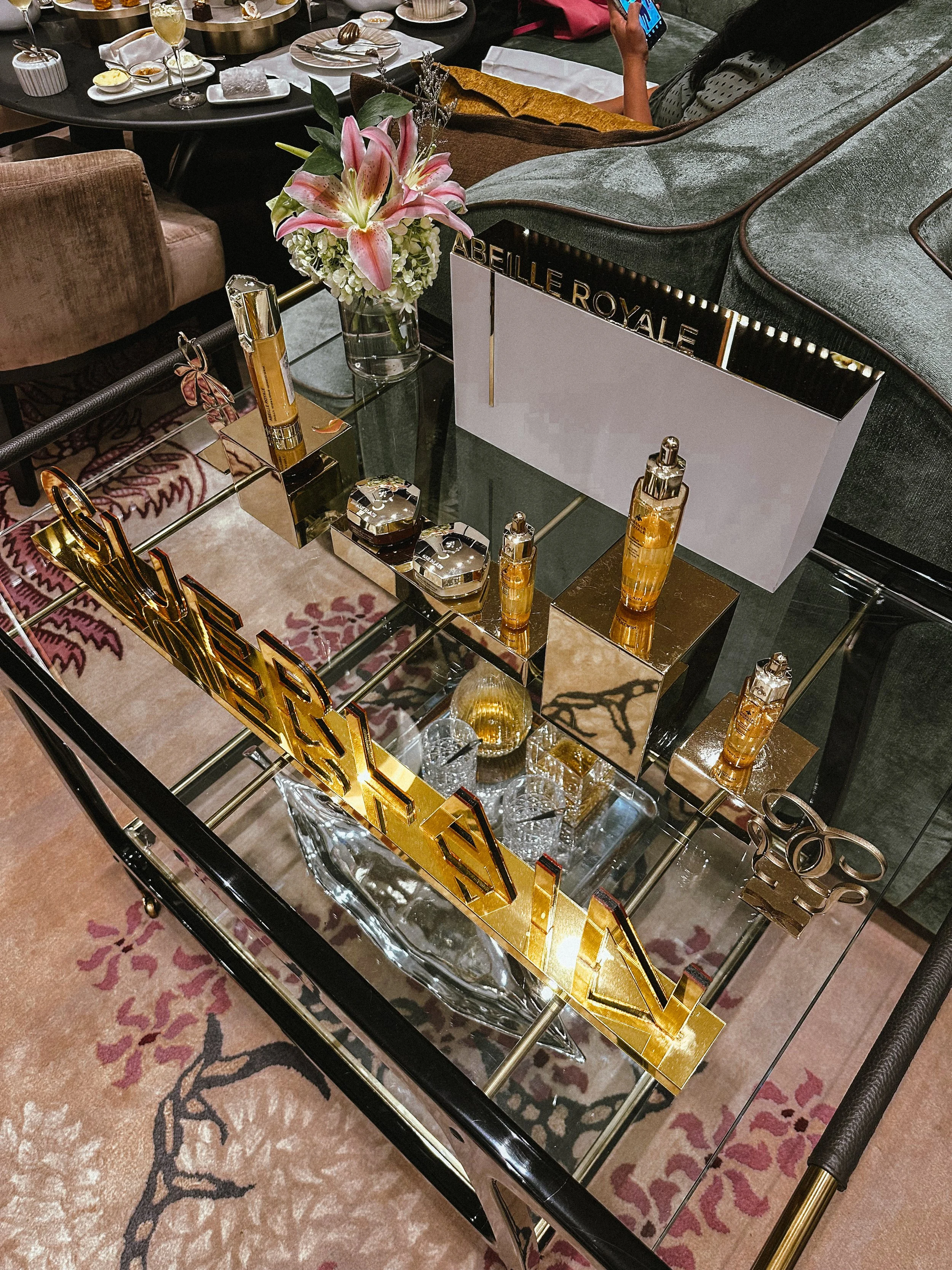 A display table featuring various skincare and beauty products, a sign with the words 'ABBEILLE ROYALE', a bouquet of pink lilies and white flowers in a glass vase, and decorative items including a mirrored block, a golden 'VOGUE' sign, and a crystal