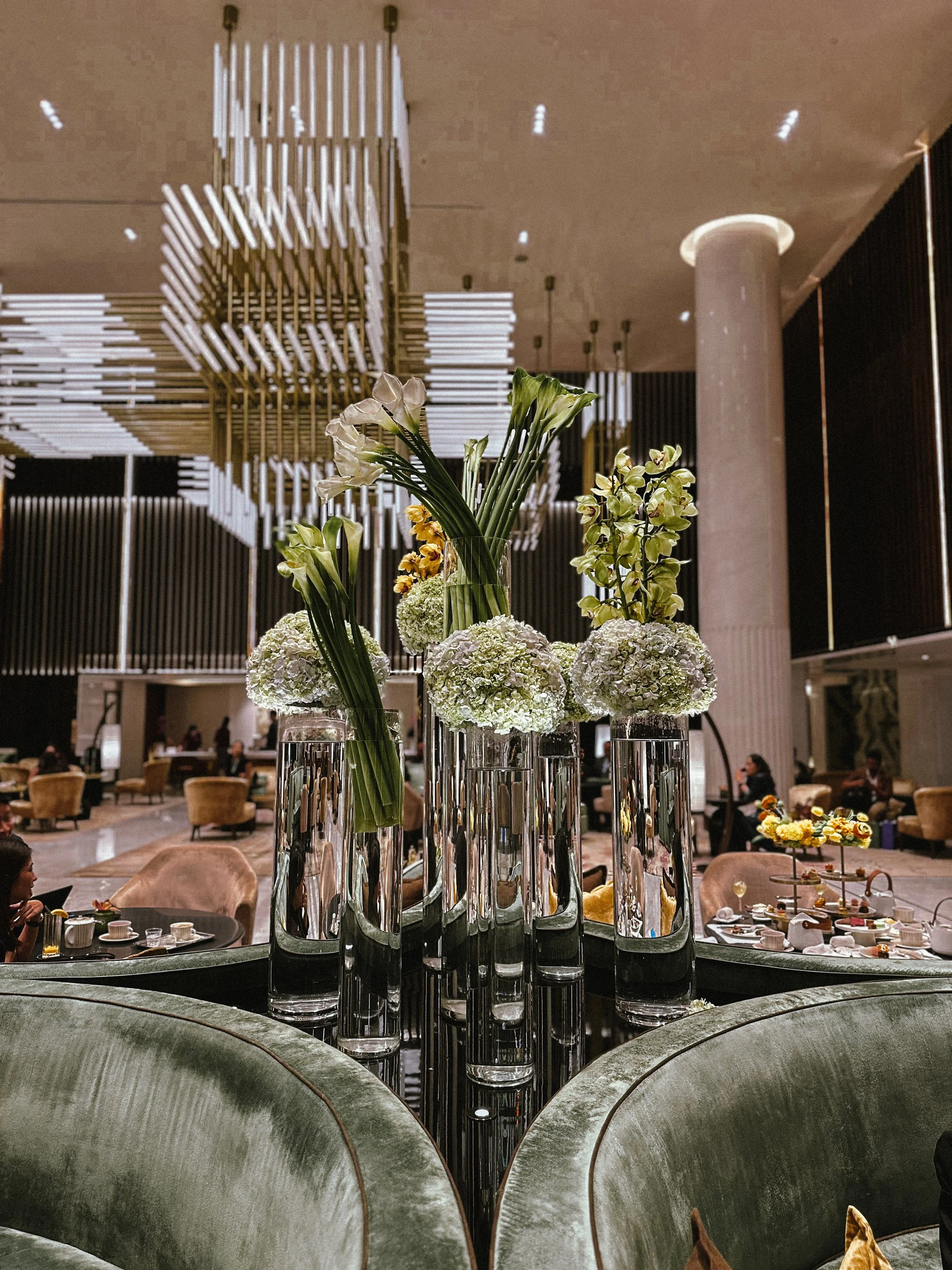 Elegant hotel lobby with tall glass vases holding white and yellow flowers on a shiny black table, with plush seating and modern lighting fixtures.