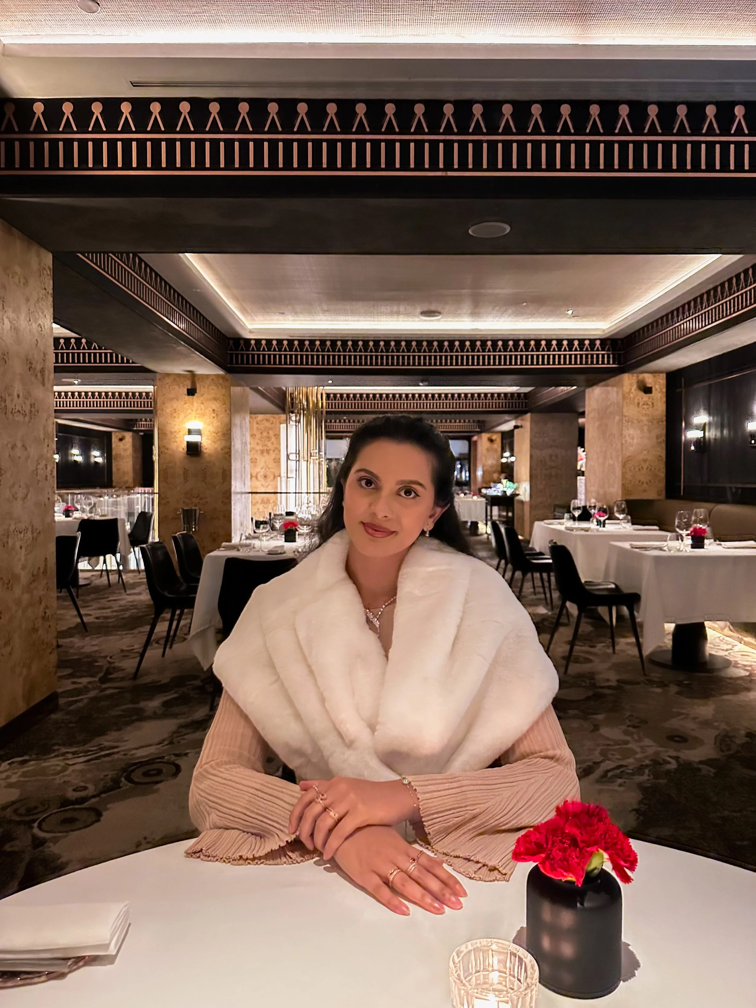 Woman sitting at a restaurant table with a white fur stole, wearing a beige dress and jewelry, with a small red flower arrangement in a black vase in front of her.