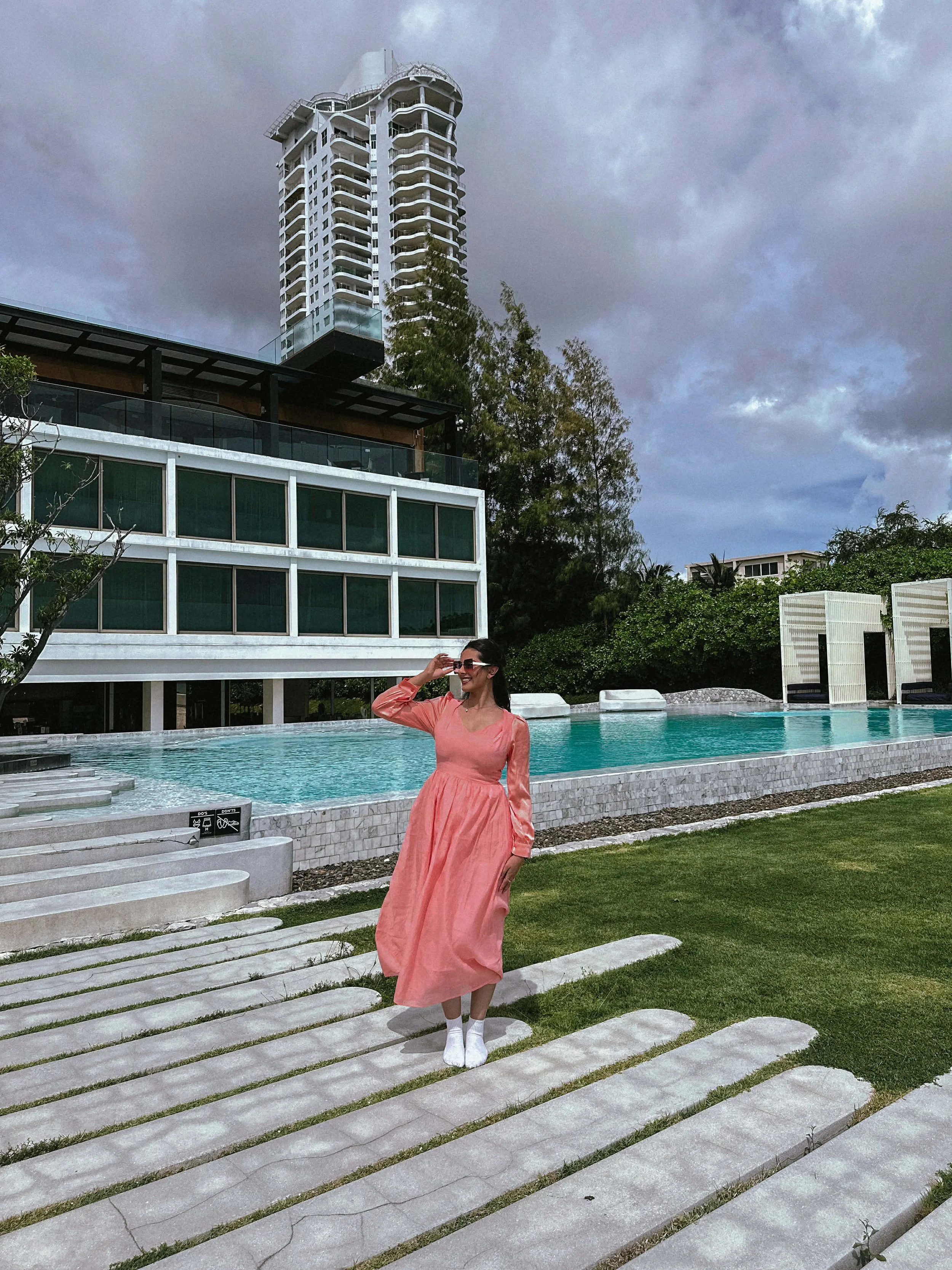 A woman in a pink dress and white shoes standing on a stone pathway by a swimming pool at a modern building with a tall, uniquely designed high-rise in the background, under cloudy skies.