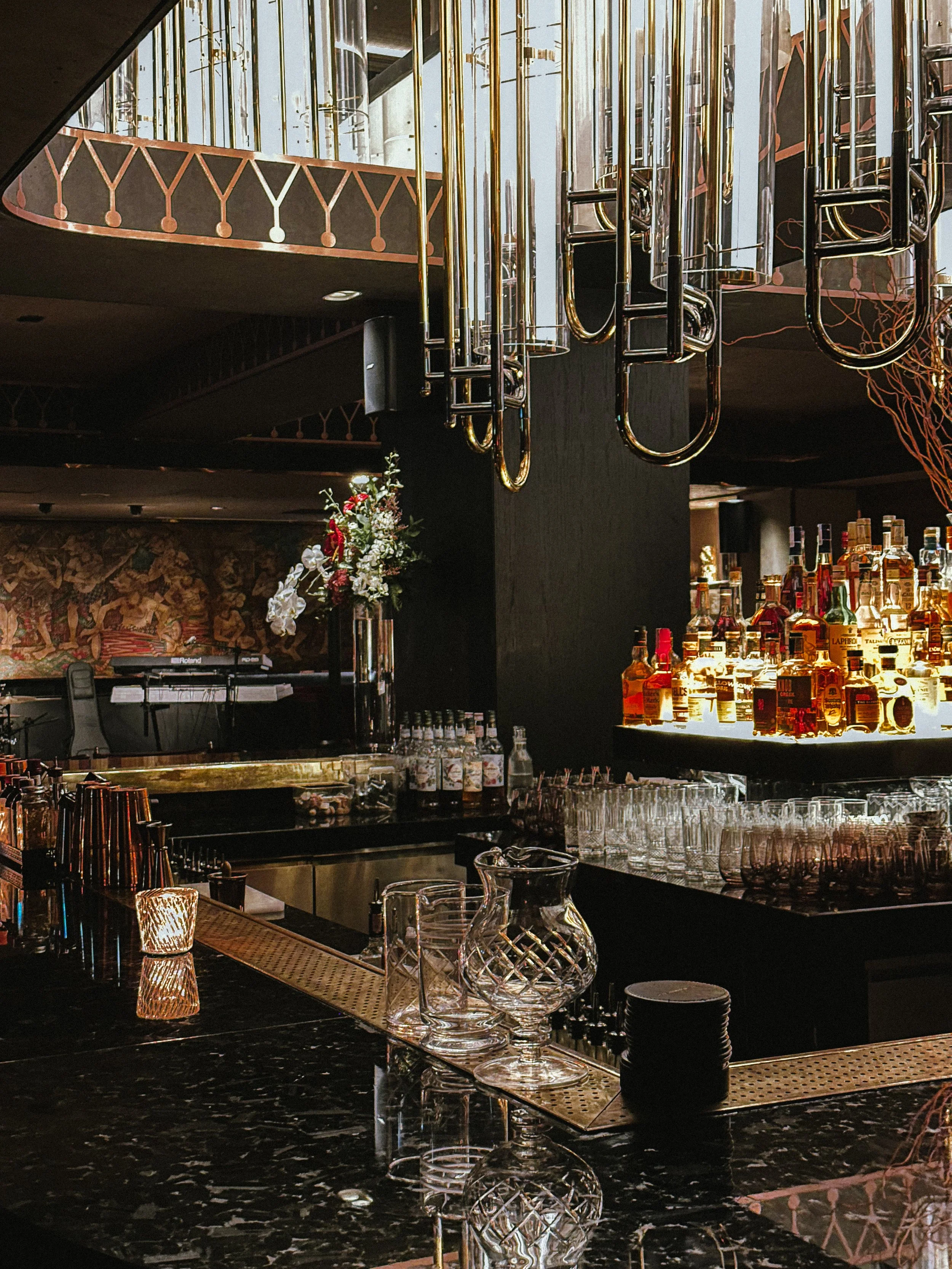 Elegant bar with a black marble counter, illuminated liquor bottles on a shelf, glassware, and a floral arrangement, with metallic hanging light fixtures.