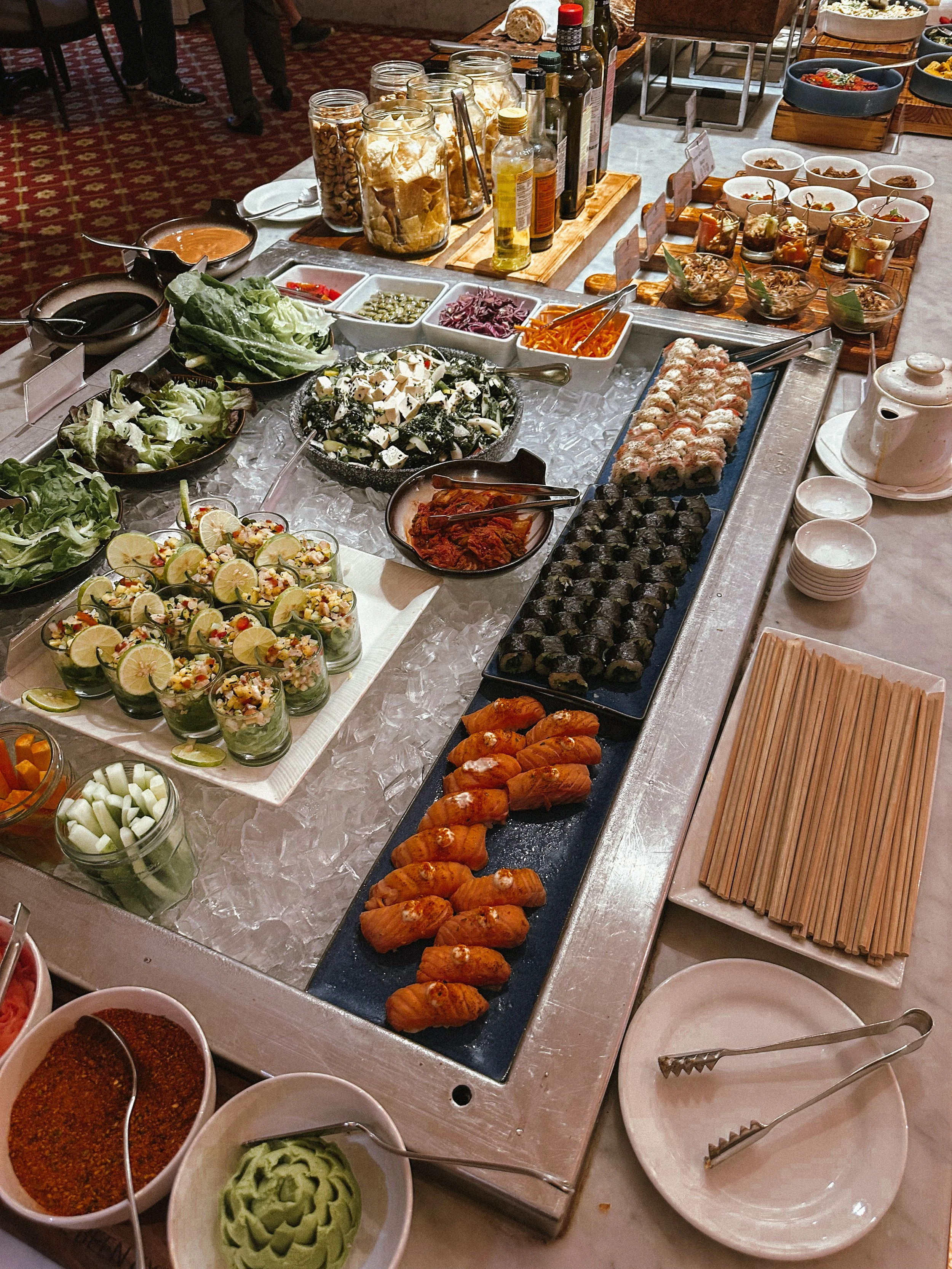A buffet table with various appetizers and ingredients including sushi rolls, salads, cucumber slices, and sauces.