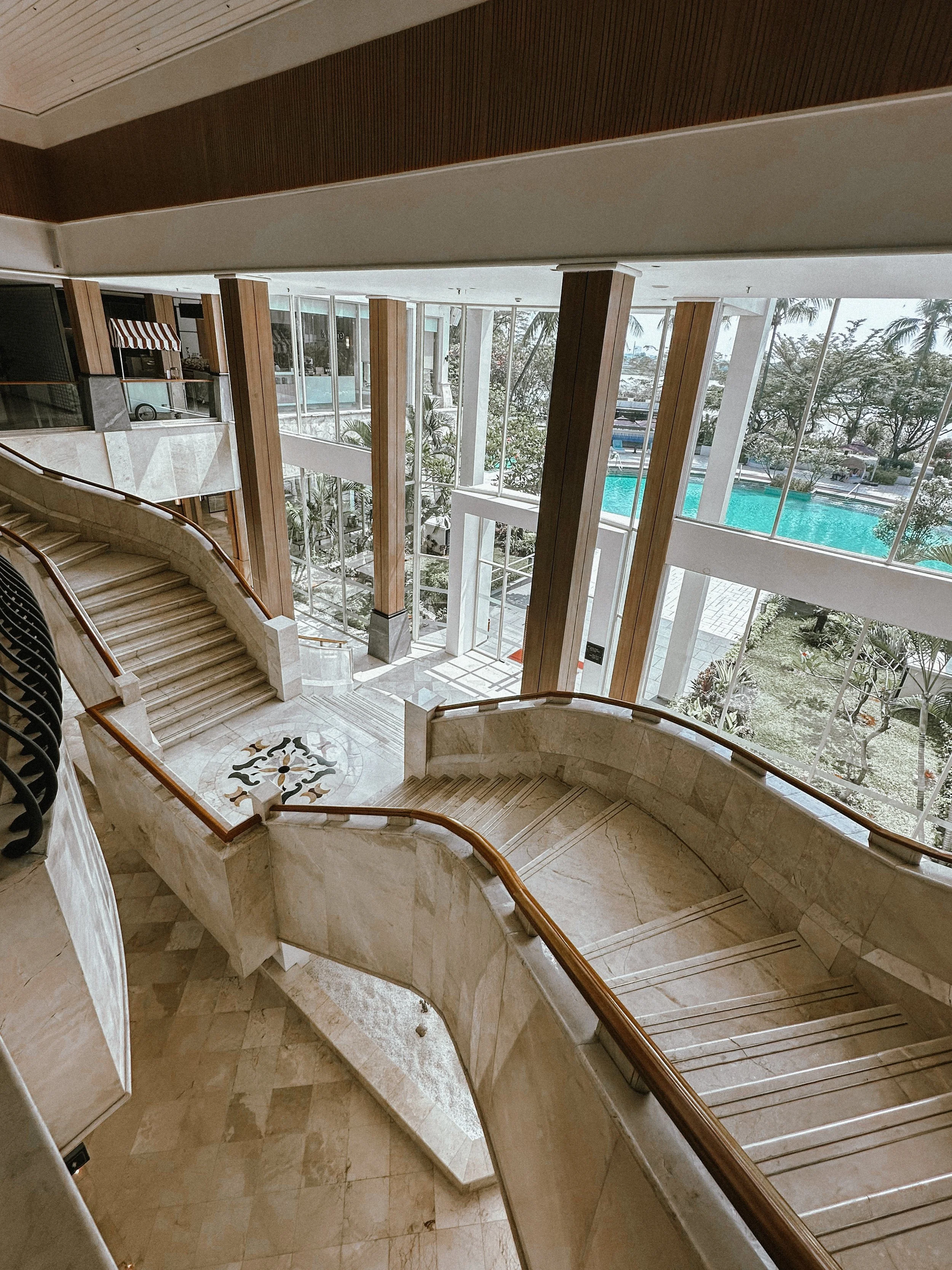 Indoor marble staircase with wooden handrails and large glass windows showing a swimming pool outside.