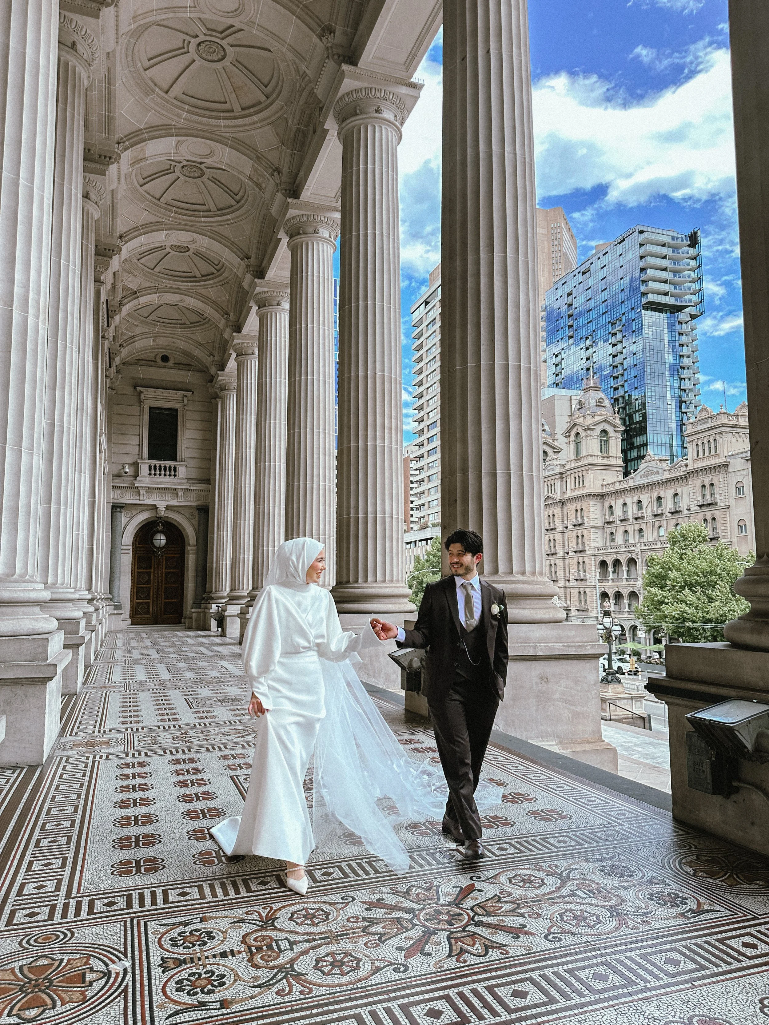 A bride in a white dress and hijab holding hands with a groom in a black suit under a grand neoclassical building with tall columns and a detailed tiled floor, overlooking a cityscape with modern skyscrapers and historic buildings.