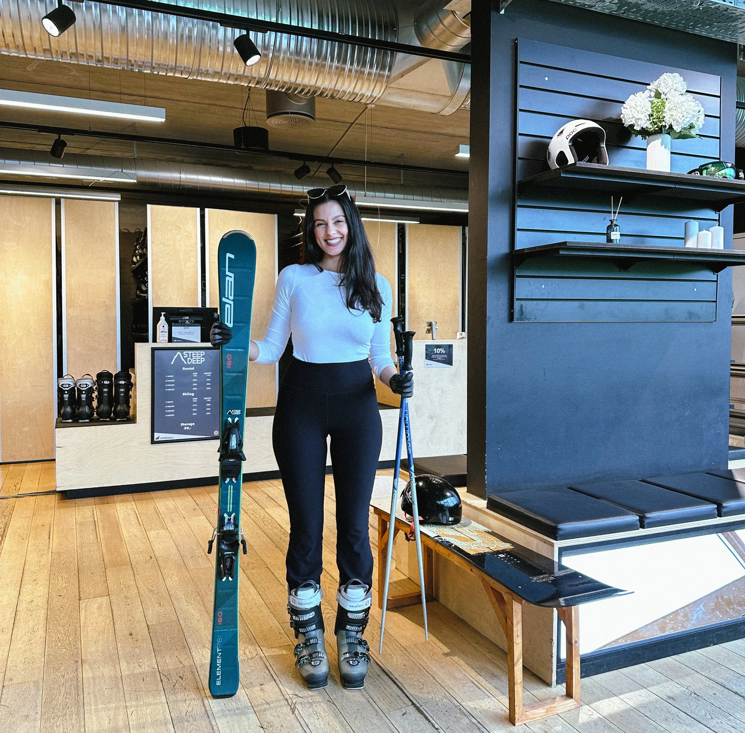 A woman smiling and holding skis and ski poles indoors at a ski rental shop.