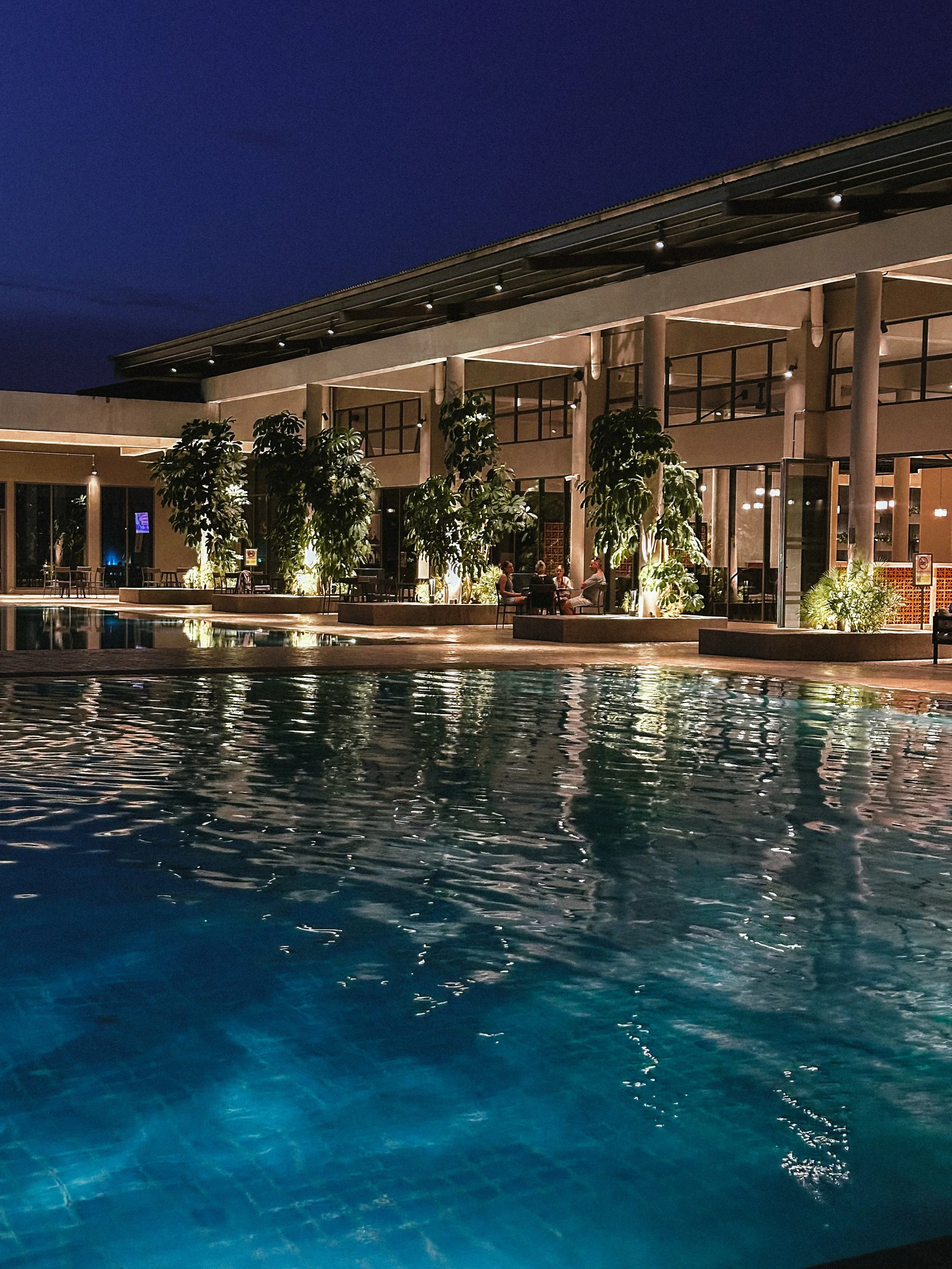 Night view of a modern hotel or resort with an outdoor swimming pool, lush green plants, and a well-lit indoor dining or lounge area visible through large glass windows.