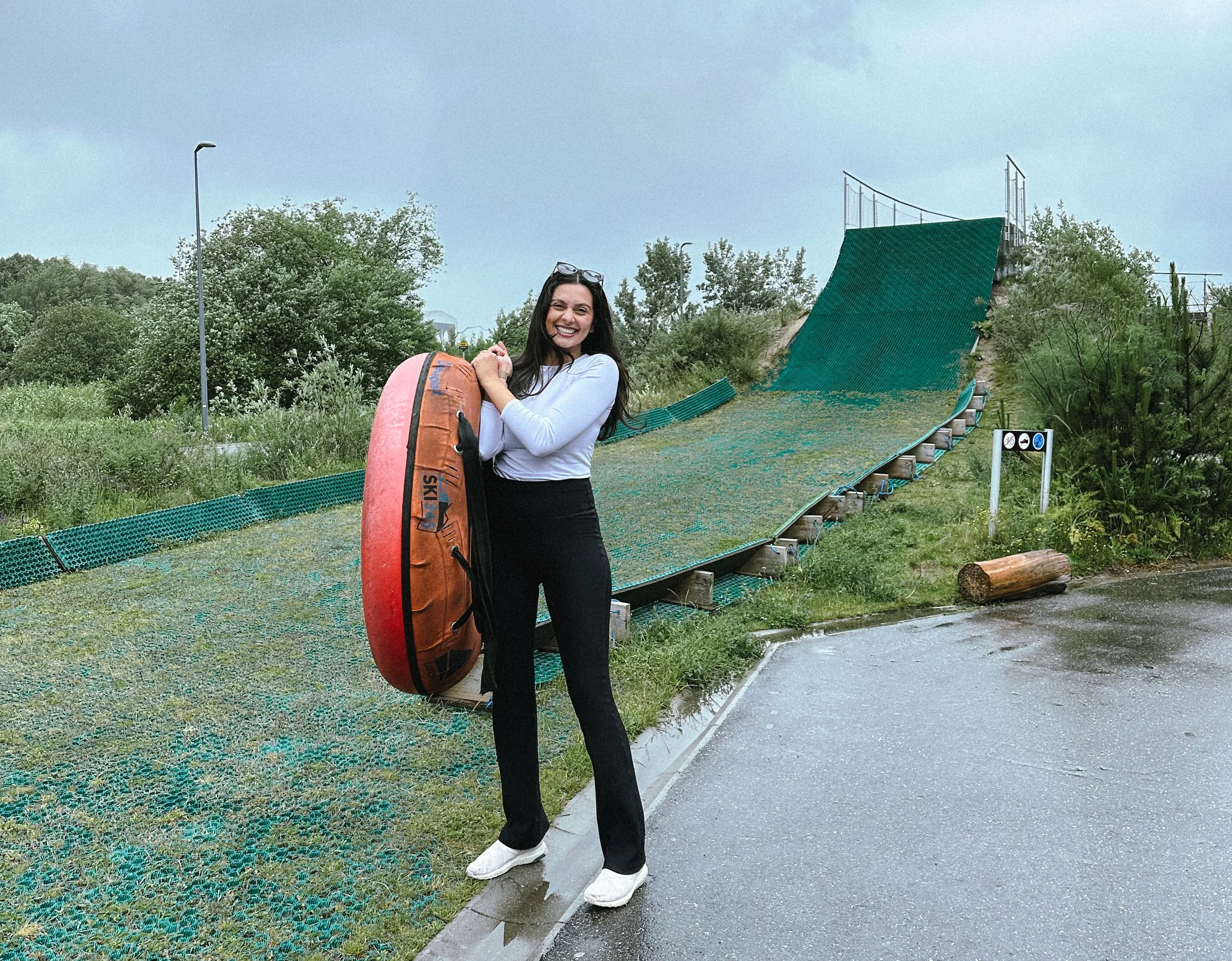 A woman standing outdoors on a wet pavement, holding a large orange and black sports bag. She is smiling and wearing a gray top, black pants, and white shoes. Behind her is a ramp with green netting, surrounded by greenery and trees.