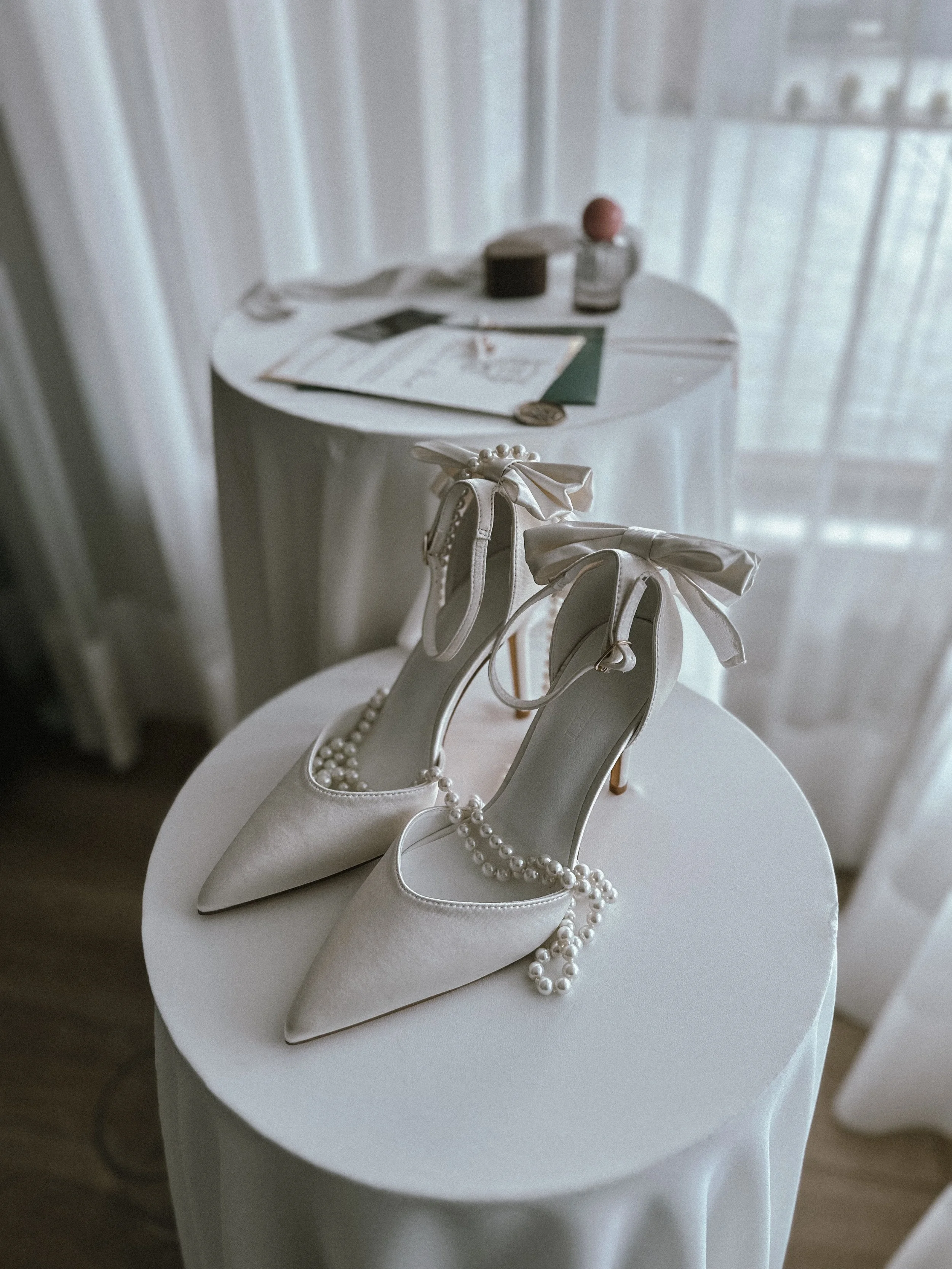 A pair of elegant white high-heeled shoes with bow and pearl embellishments resting on a round table. The shoes are adorned with pearl necklaces draped over them. In the background, there is a table with a playlist, a glass jar, and a candle, with sh
