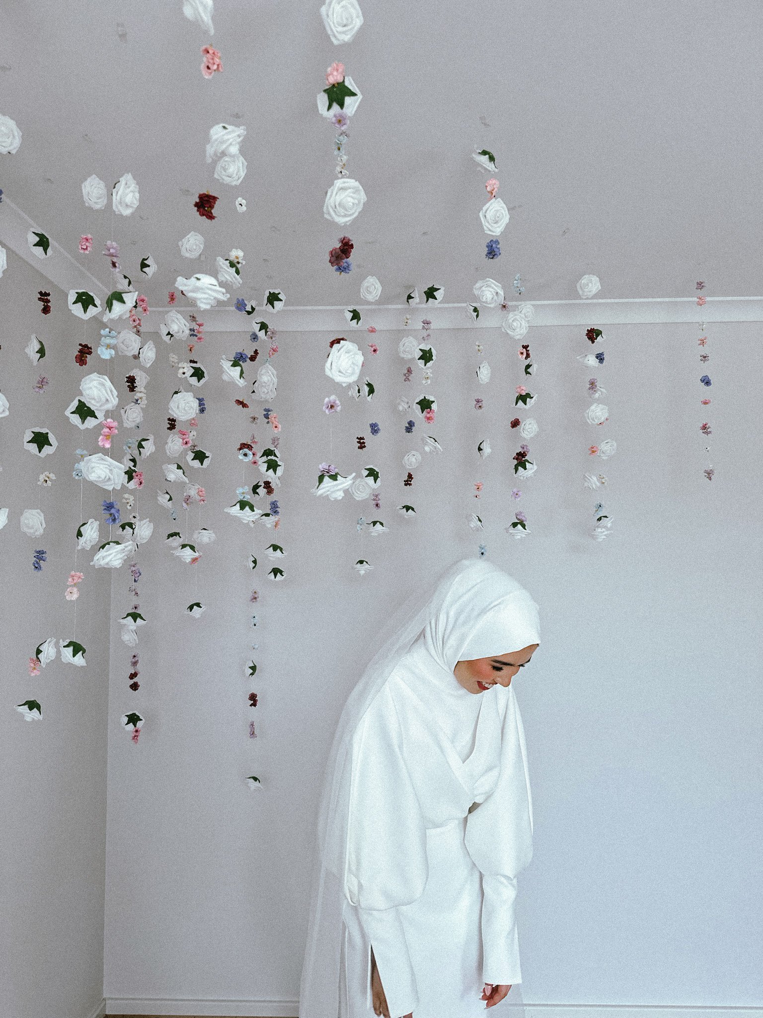 A woman in a white hijab and dress bowing under hanging floral decorations on a white ceiling.