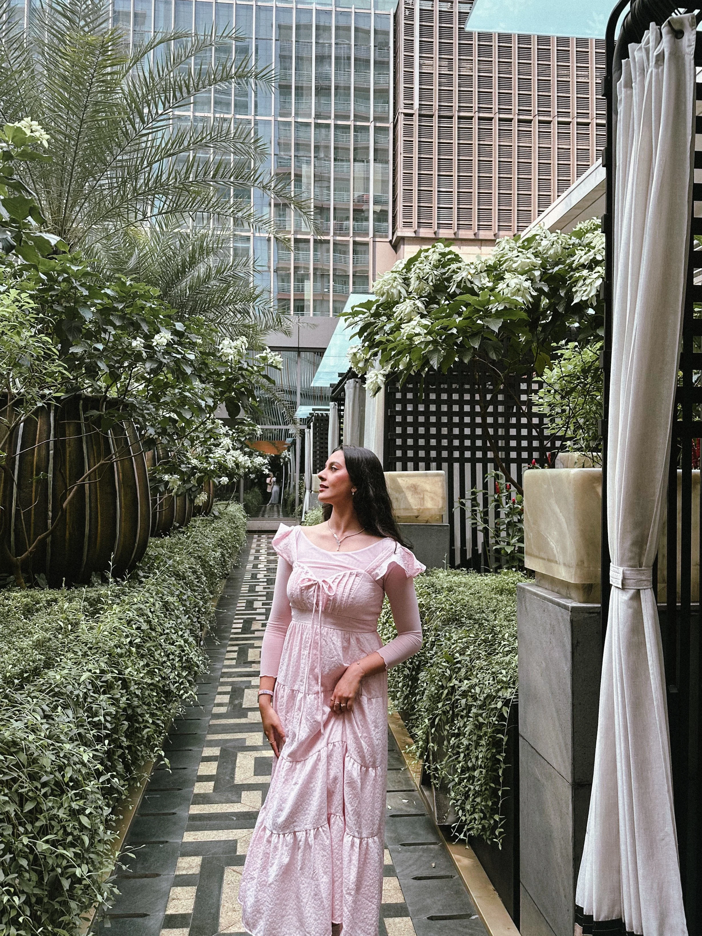 A woman stands on a patterned outdoor pathway surrounded by greenery and potted plants in an urban setting with tall buildings in the background.