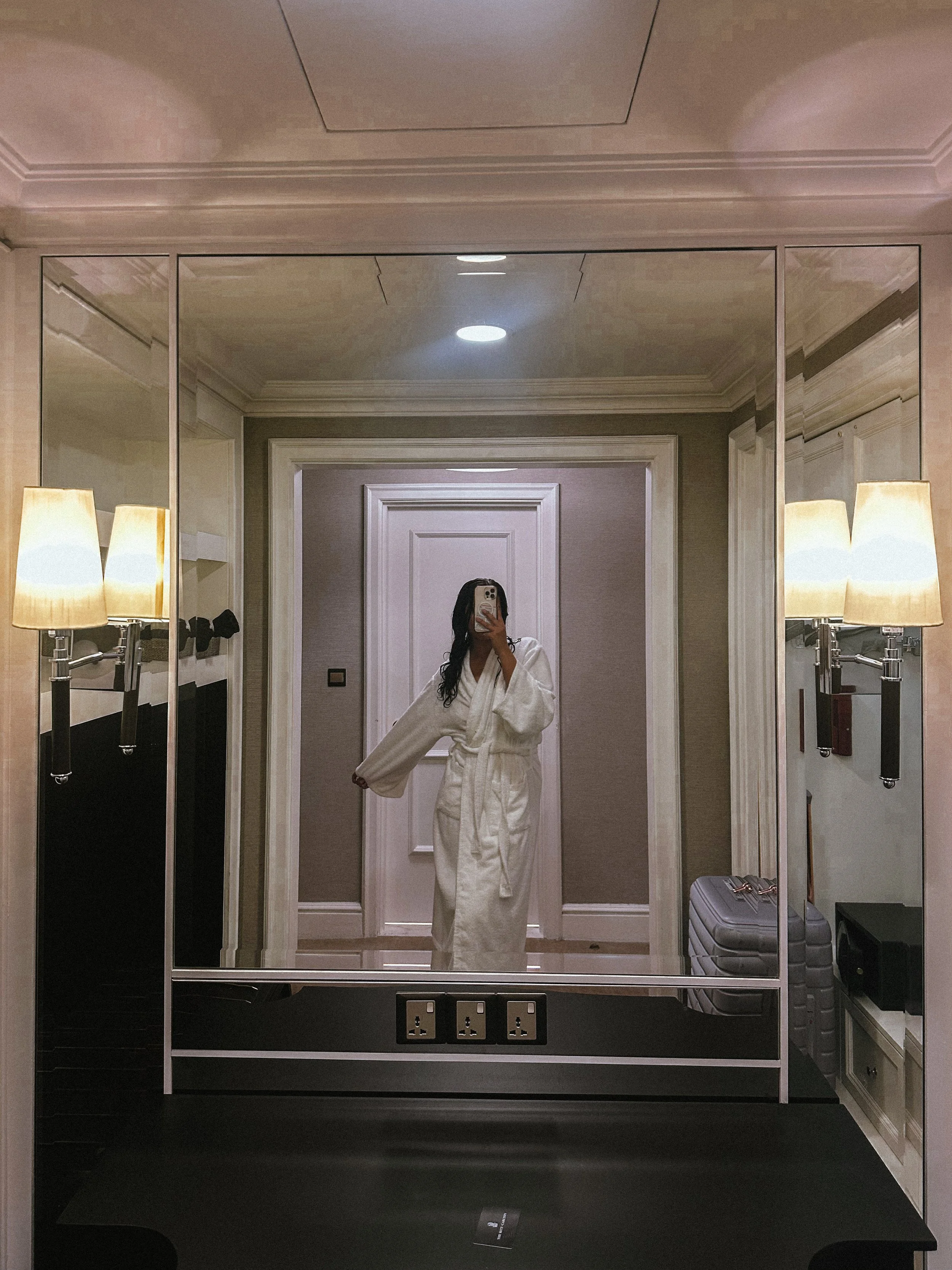 Reflection of a woman in a large mirror taking a photo with her phone, dressed in a white bathrobe, in a hotel room with beige walls, lamps on each side of the mirror, and a suitcase on the right side.