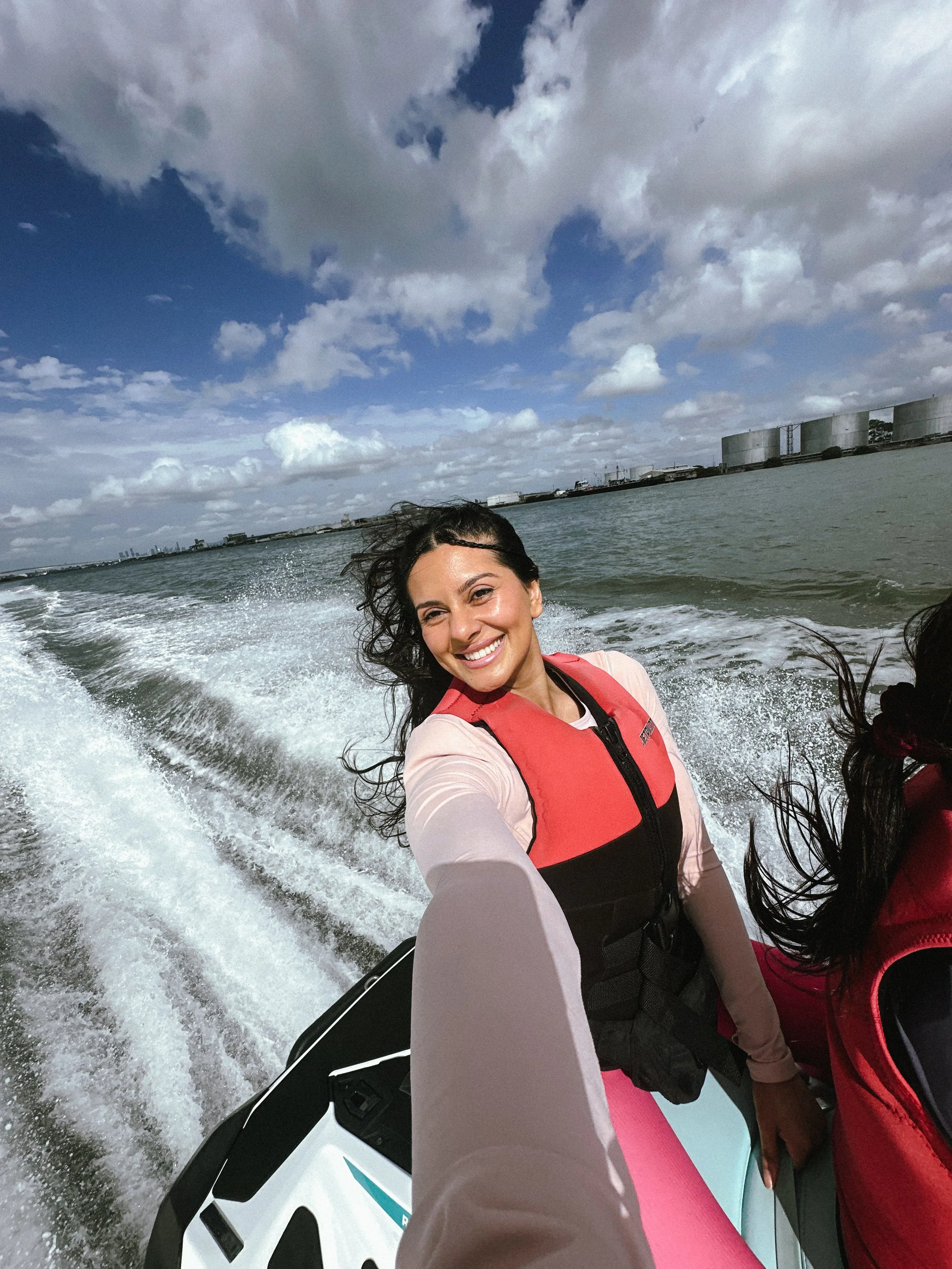 A woman smiling on a boat ride wearing a red life jacket, with water and a partly cloudy sky in the background.