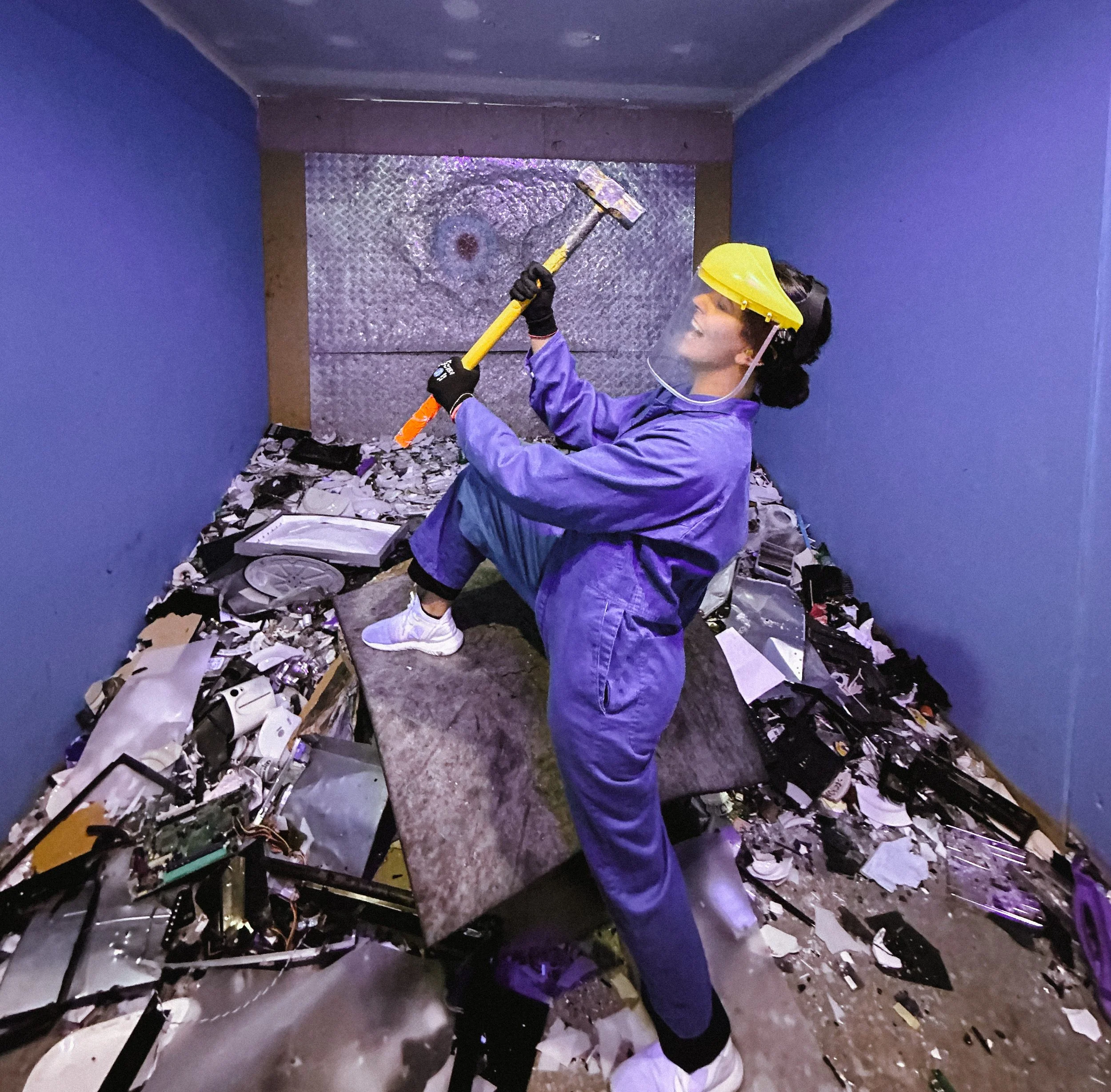 Woman in blue coveralls and protective gear smashing electronic waste with a sledgehammer in a small enclosed space.