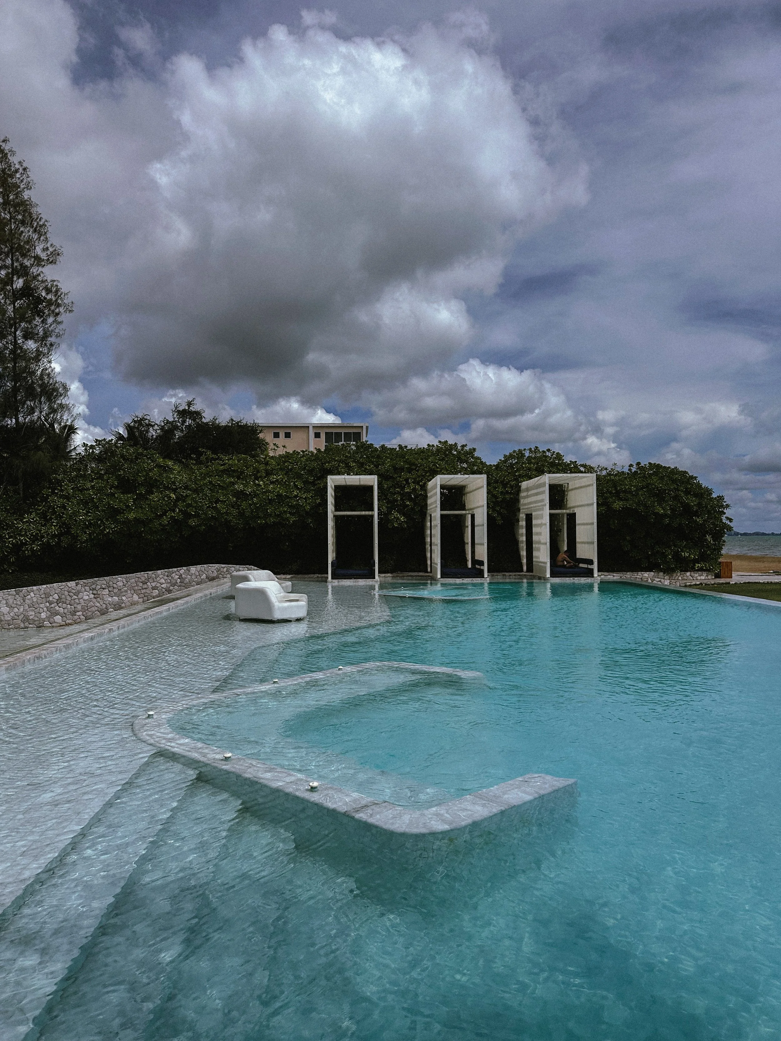 A swimming pool with three white cabanas and lounge chairs under cloudy skies, with greenery and a building in the background.