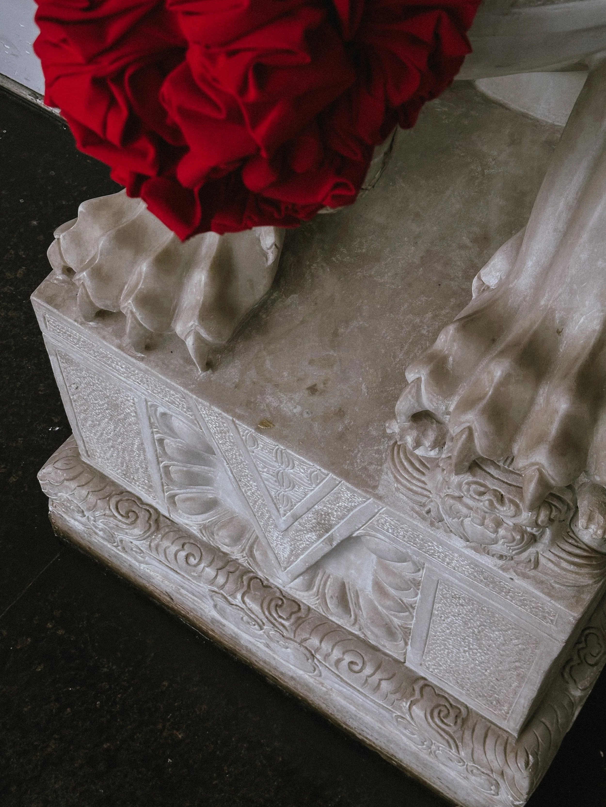 Close-up of a carved stone or marble pedestal with ornate details, supporting a statue draped in red cloth.