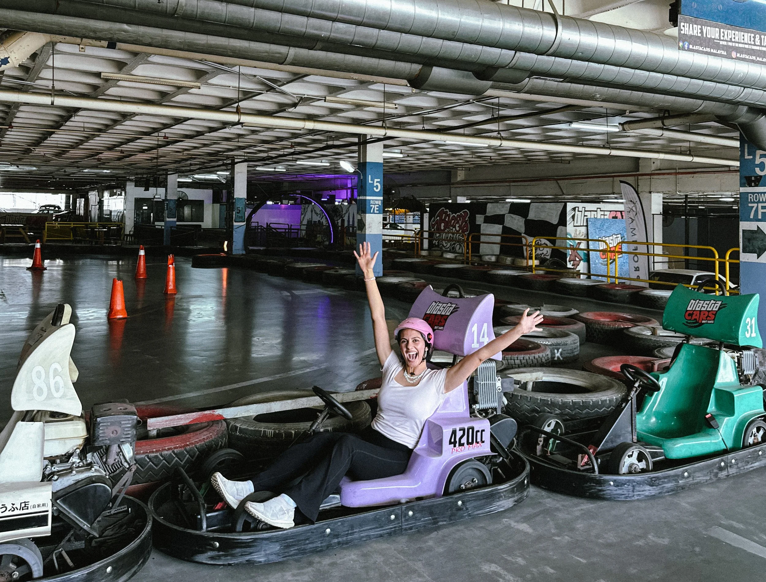 A woman sitting in a go-kart in an indoor go-kart racing facility, smiling and raising her arms in excitement, surrounded by other colorful go-karts and track barriers.