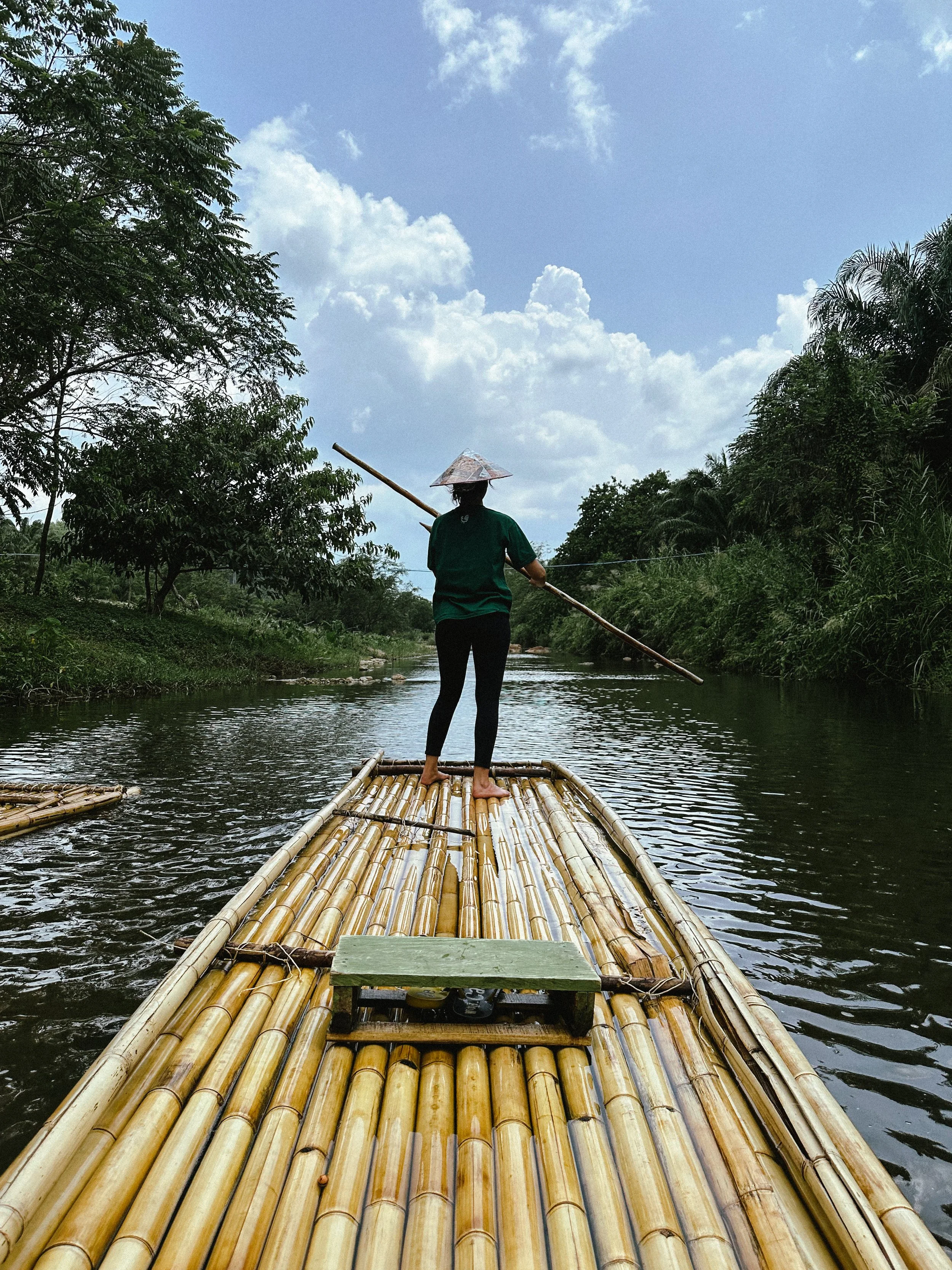 A person wearing a conical Asian hat standing on a bamboo raft, navigating a river with a pole, surrounded by lush greenery and blue sky with clouds.