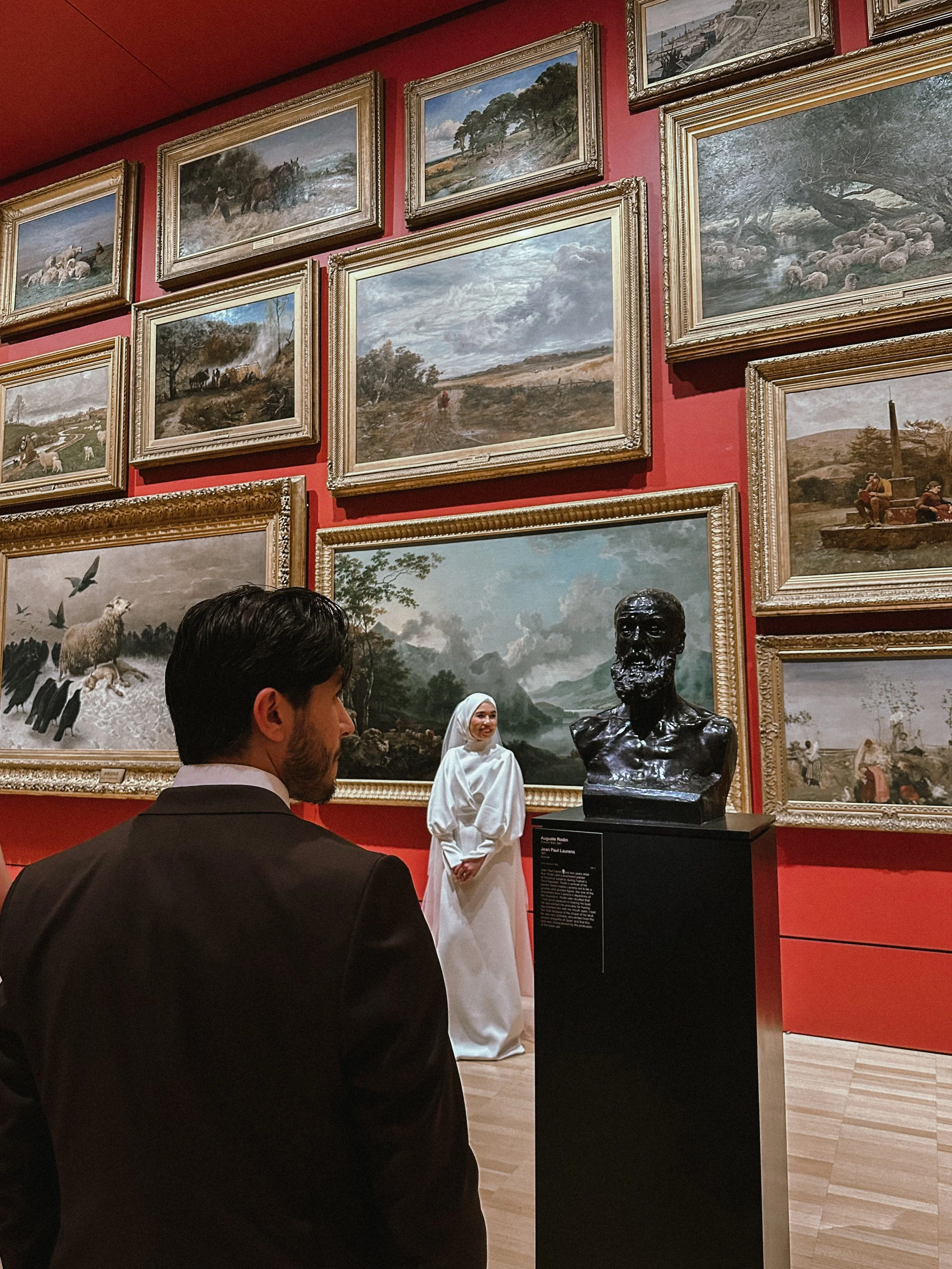 People viewing a black bust sculpture of a man in an art gallery, with framed landscape paintings on the red wall behind them.