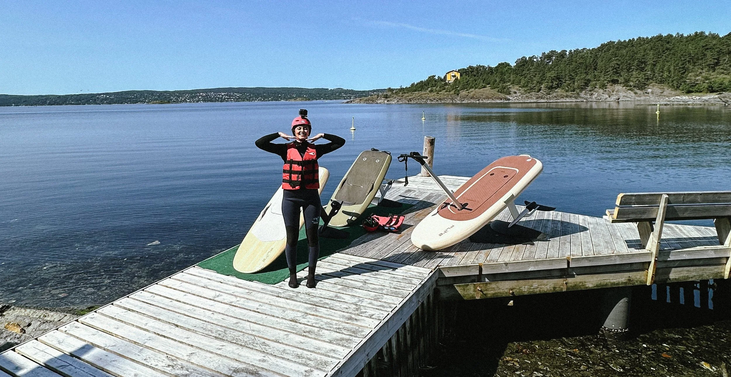 A person wearing a life jacket and helmet standing on a dock near water, with wakeboards and a paddleboard nearby, and a scenic lake with trees and hills in the background.