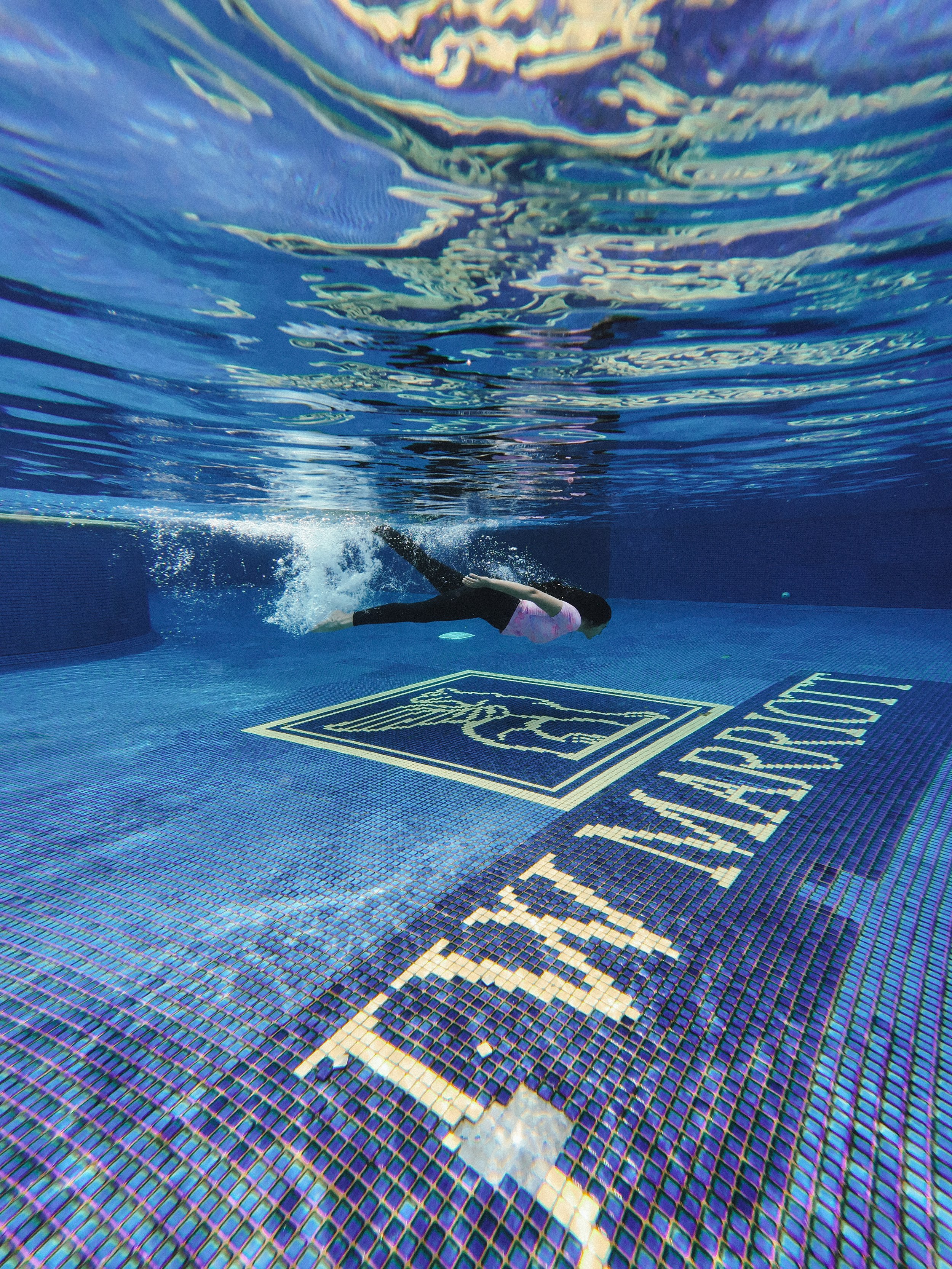 A person swimming underwater in a pool with a logo and white text on the pool floor.