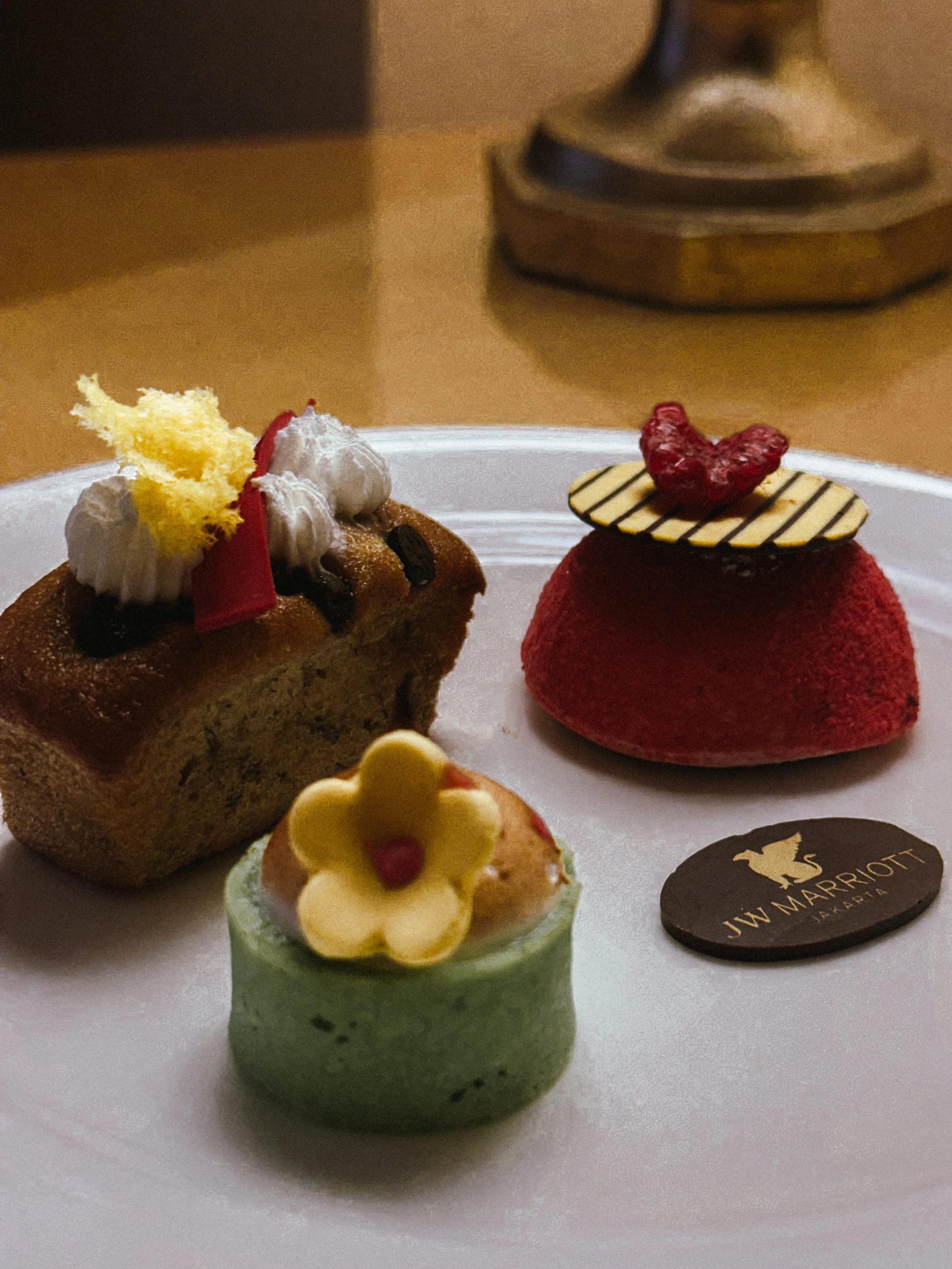 Assorted desserts on a white plate including a chocolate chip bread, a green mousse cake with a yellow flower-shaped decoration, a red dome-shaped mousse with a striped chocolate disc and a red heart garnish, and a dark oval chocolate piece with a go