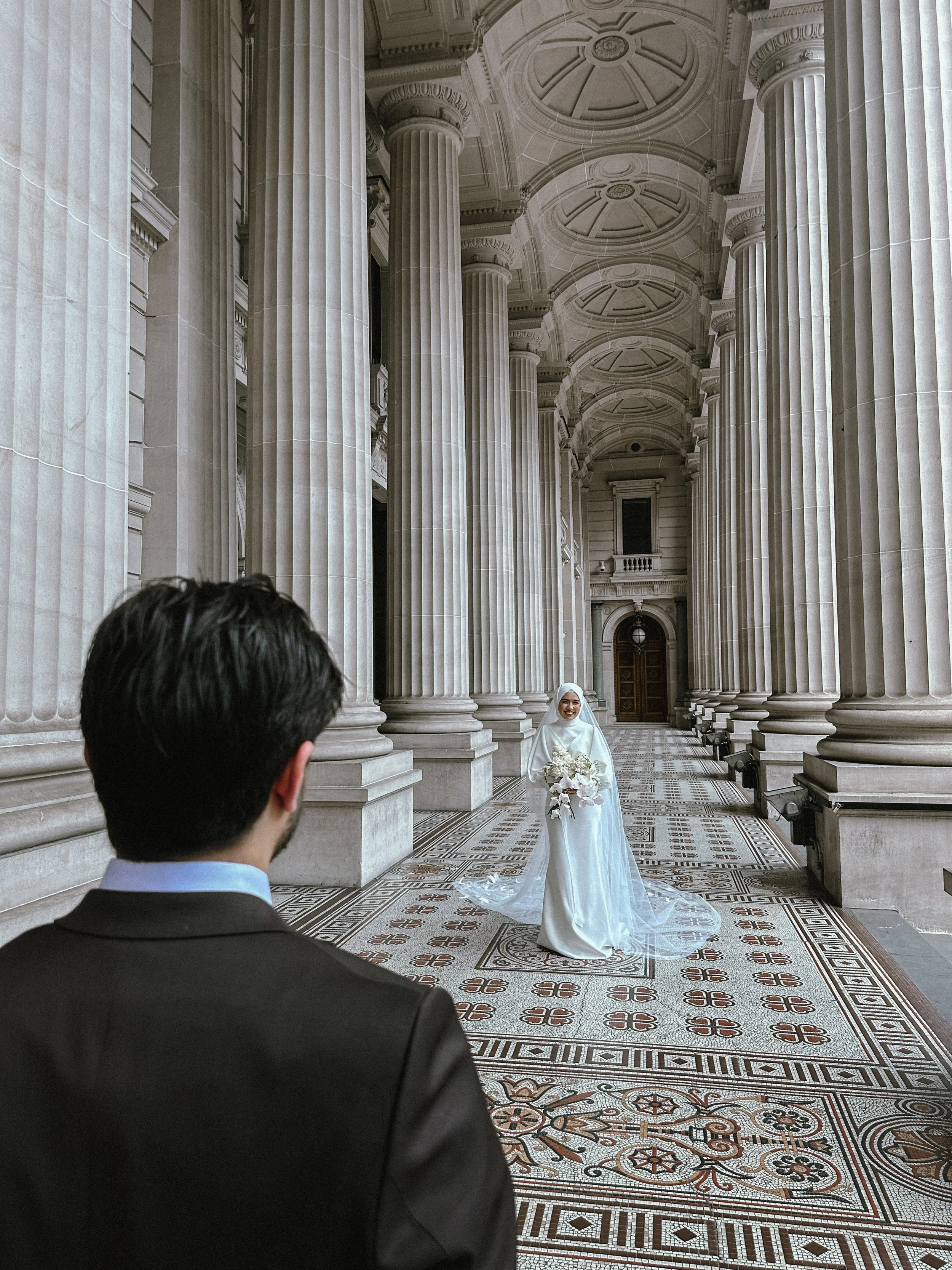 A bride in a white gown holding a bouquet of flowers, standing in a grand hall with tall columns and intricate ceiling designs, while a groom in a dark suit looks at her.