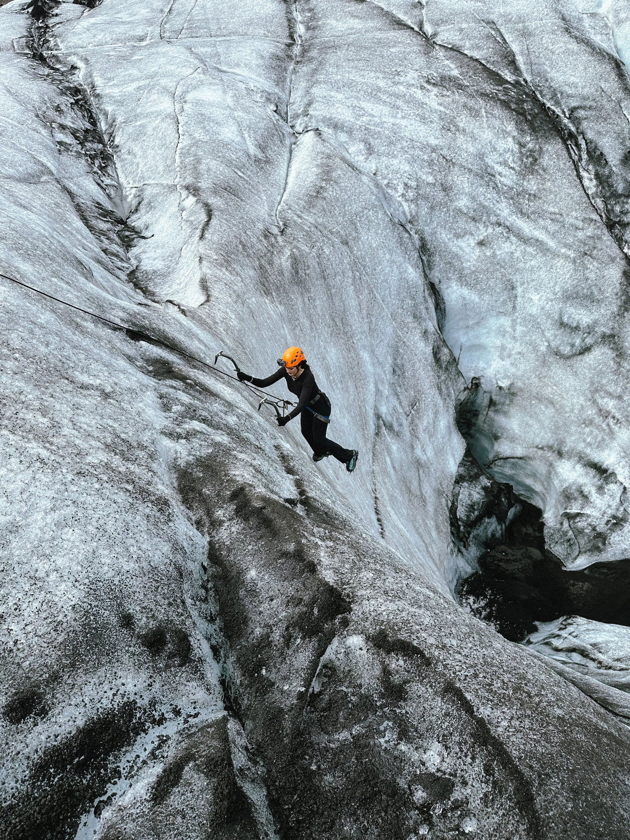 A person in black climbing icy rock with safety gear and orange helmet.
