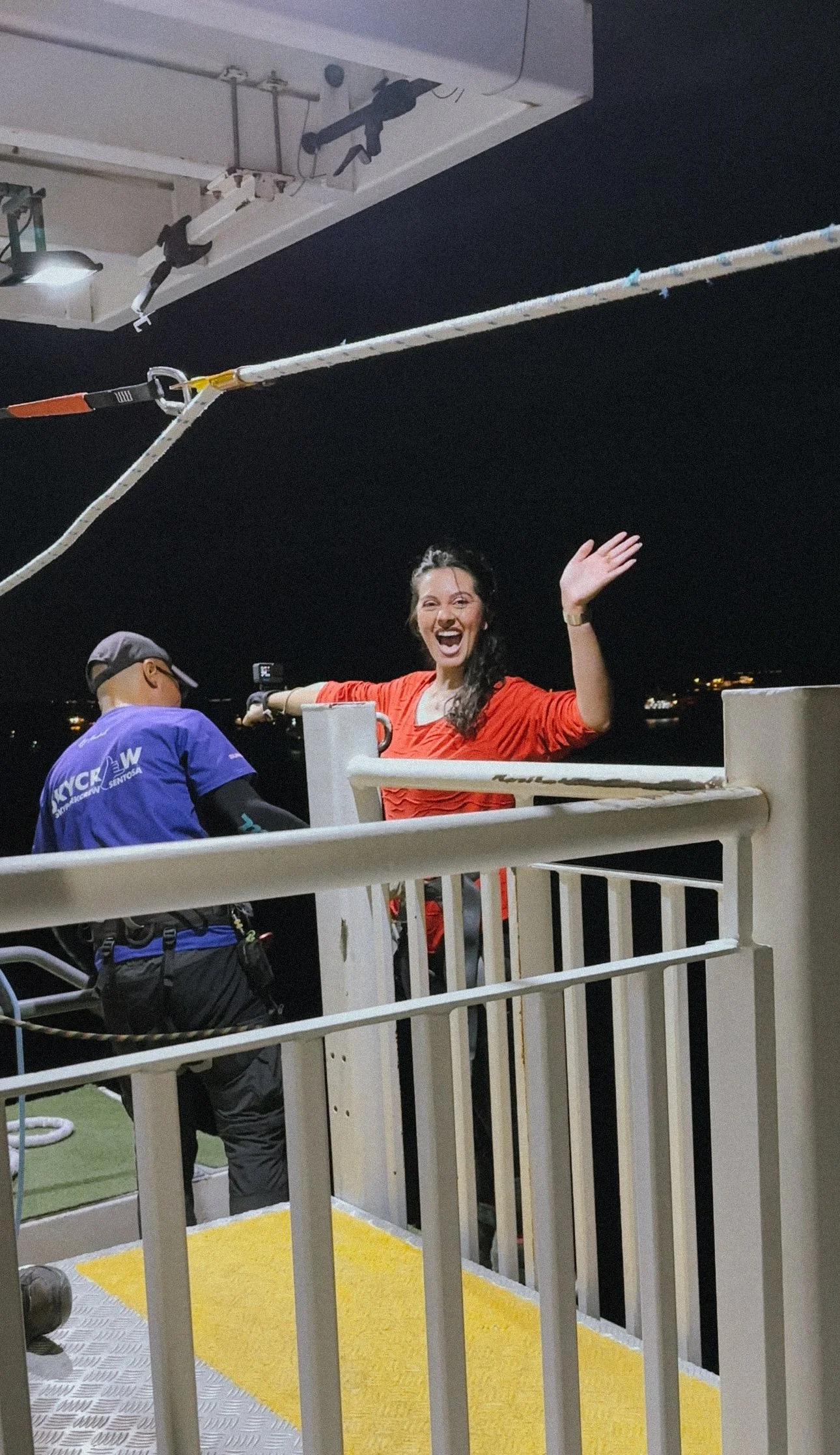 A woman on a boat deck at night, smiling and waving at the camera. She is wearing a red shirt. There is a person in a blue shirt facing away, possibly working or taking a picture. The boat's safety railings and ropes are visible, with a dark night sk