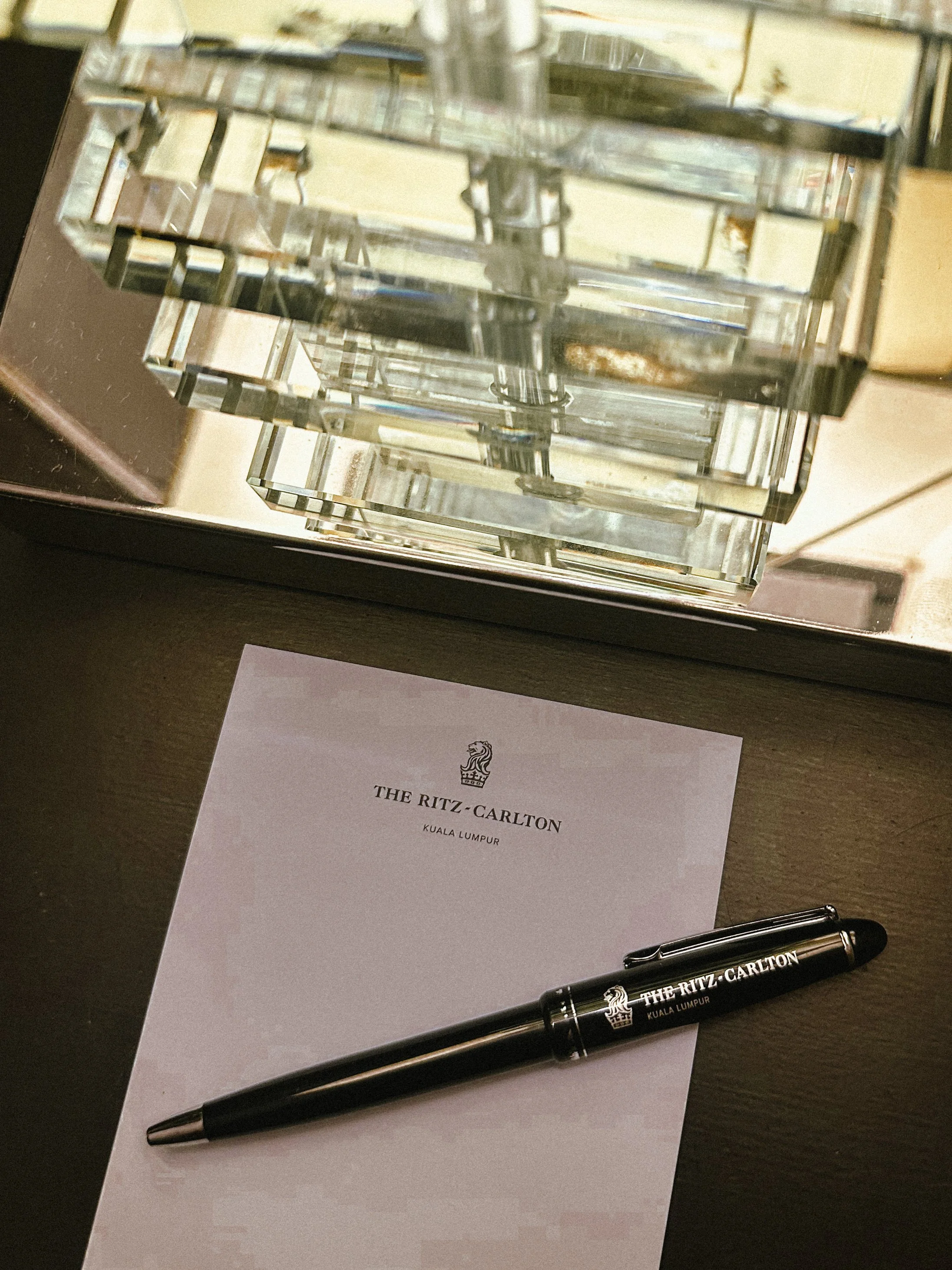 A black pen with the logo and name of The Ritz-Carlton hotel, placed on a white paper with hotel details, beside a glass sculptural decorative piece on a reflective surface.