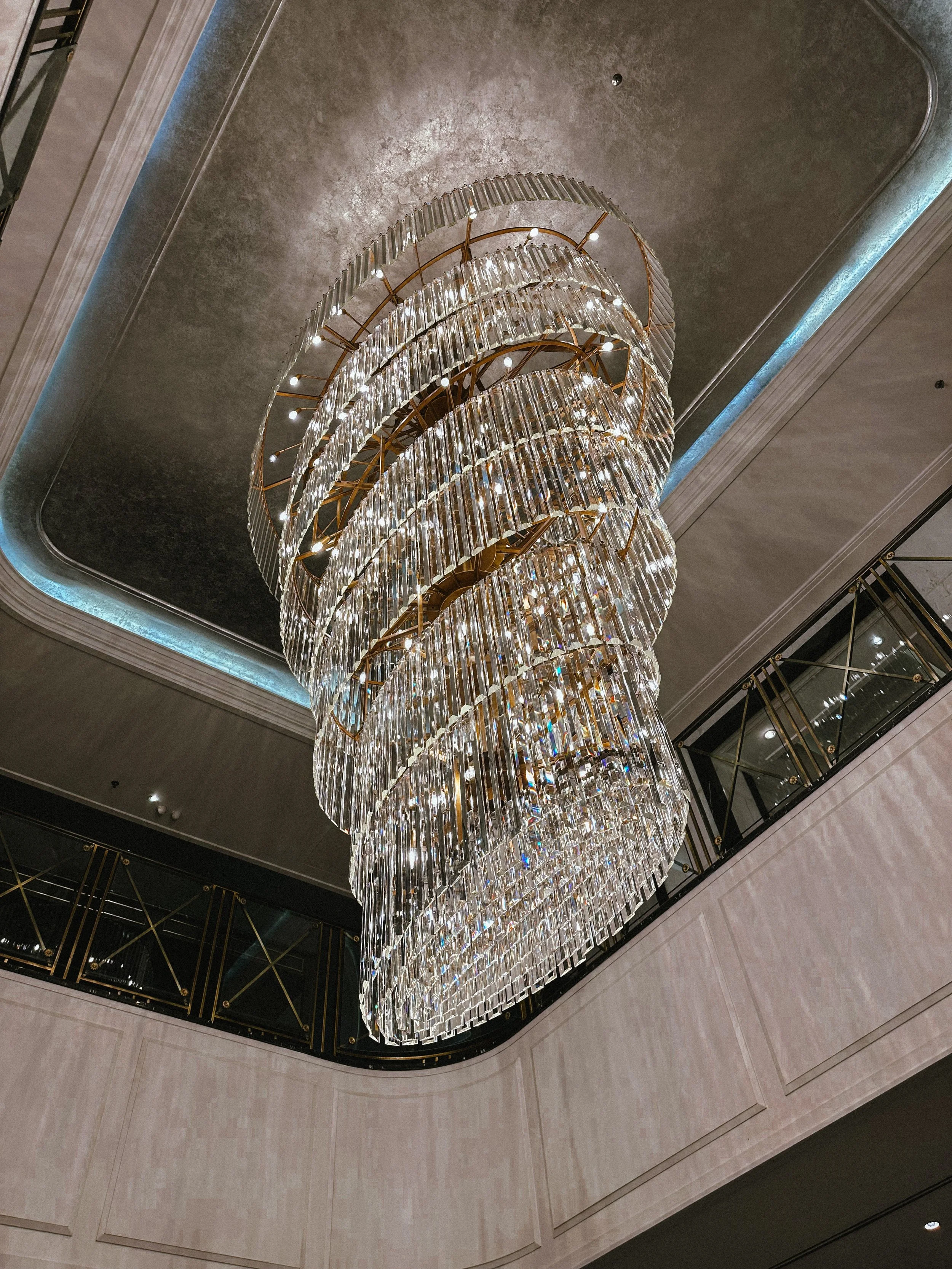 A large, decorative chandelier made of multiple tiers of glass and metal hangs from the ceiling in a hotel or luxury building lobby.