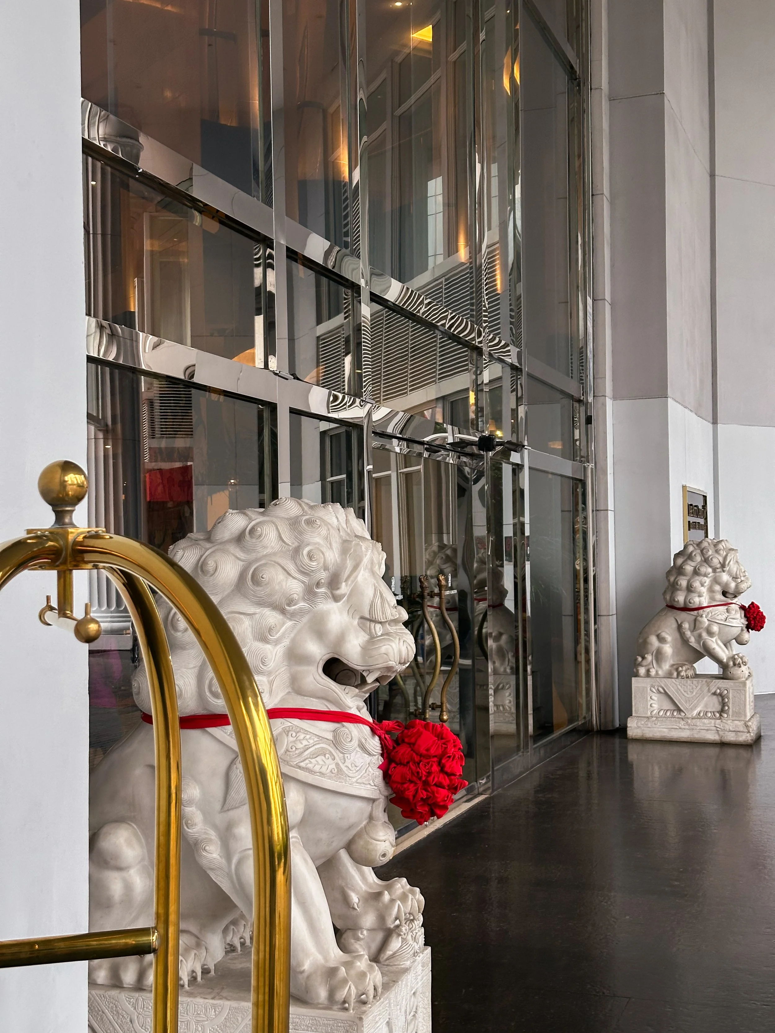 Two white stone lion statues with red flower garlands, one near a gold luggage cart and the other further down the corridor, with a building entrance and glass windows reflecting the interior.