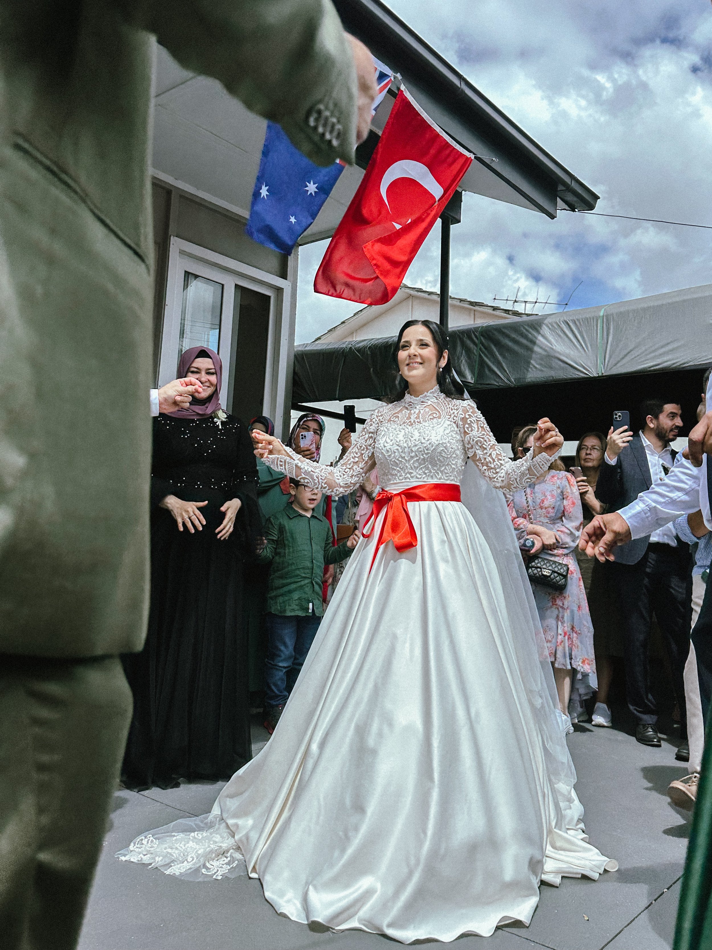 A woman in a white wedding dress with lace detail and a red sash dances outdoors, surrounded by onlookers. Flags of Australia, Turkey, and another country hang above.