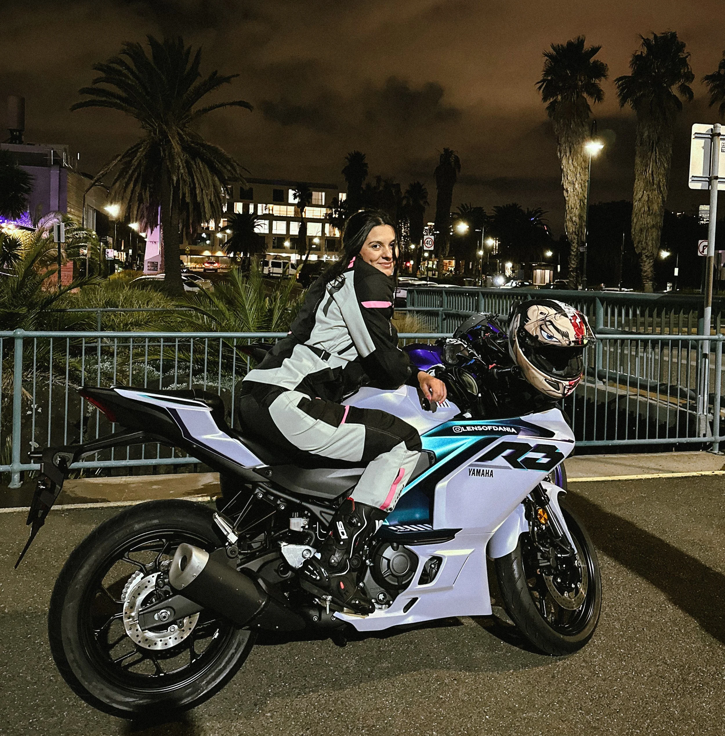 A woman in motorcycle gear sitting on a Yamaha sportbike at night, with a helmet resting on the bike, in an urban area with palm trees and streetlights.