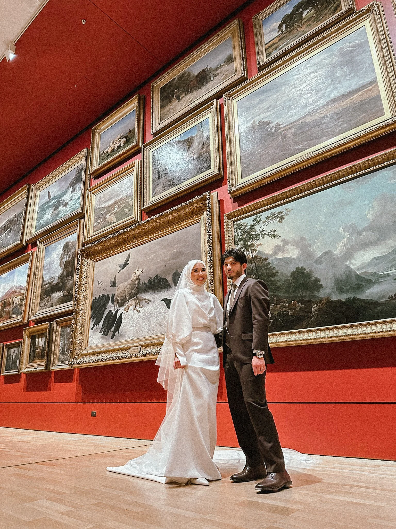 A bride and groom stand together in an art museum with many framed landscape paintings on a red wall behind them.