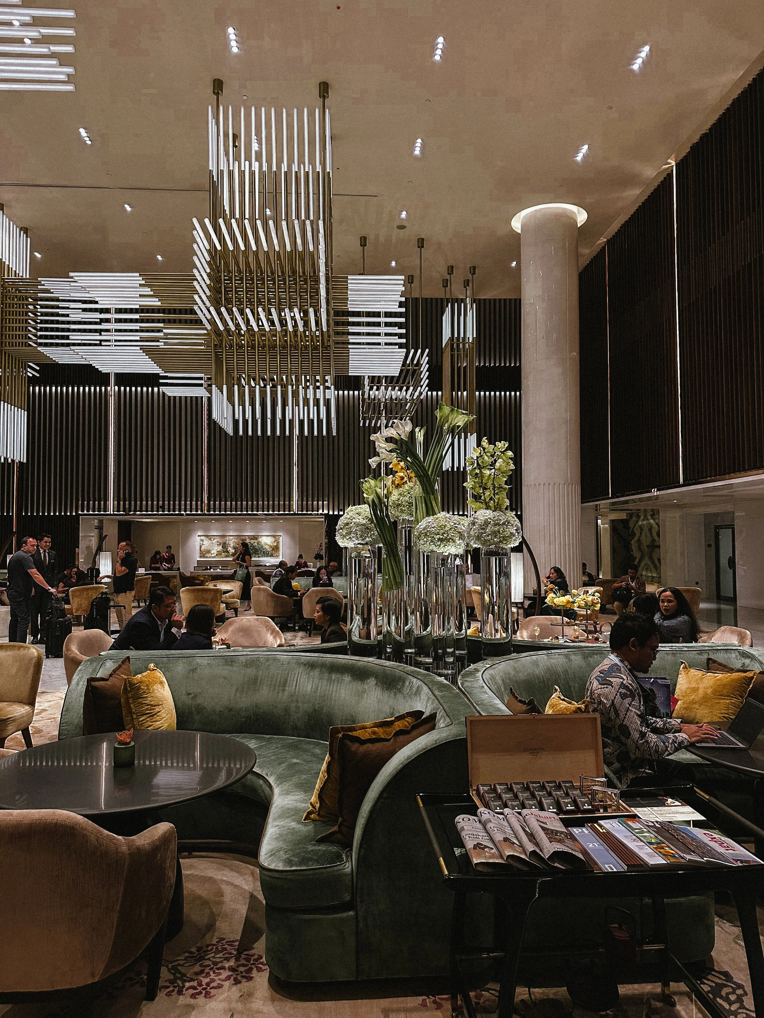 A luxurious hotel lobby with high ceiling, modern chandeliers, and elegant furniture including a curved green velvet sofa with pillows, flower arrangements in tall vases, and people seated and working. There are staff and other guests in the backgrou