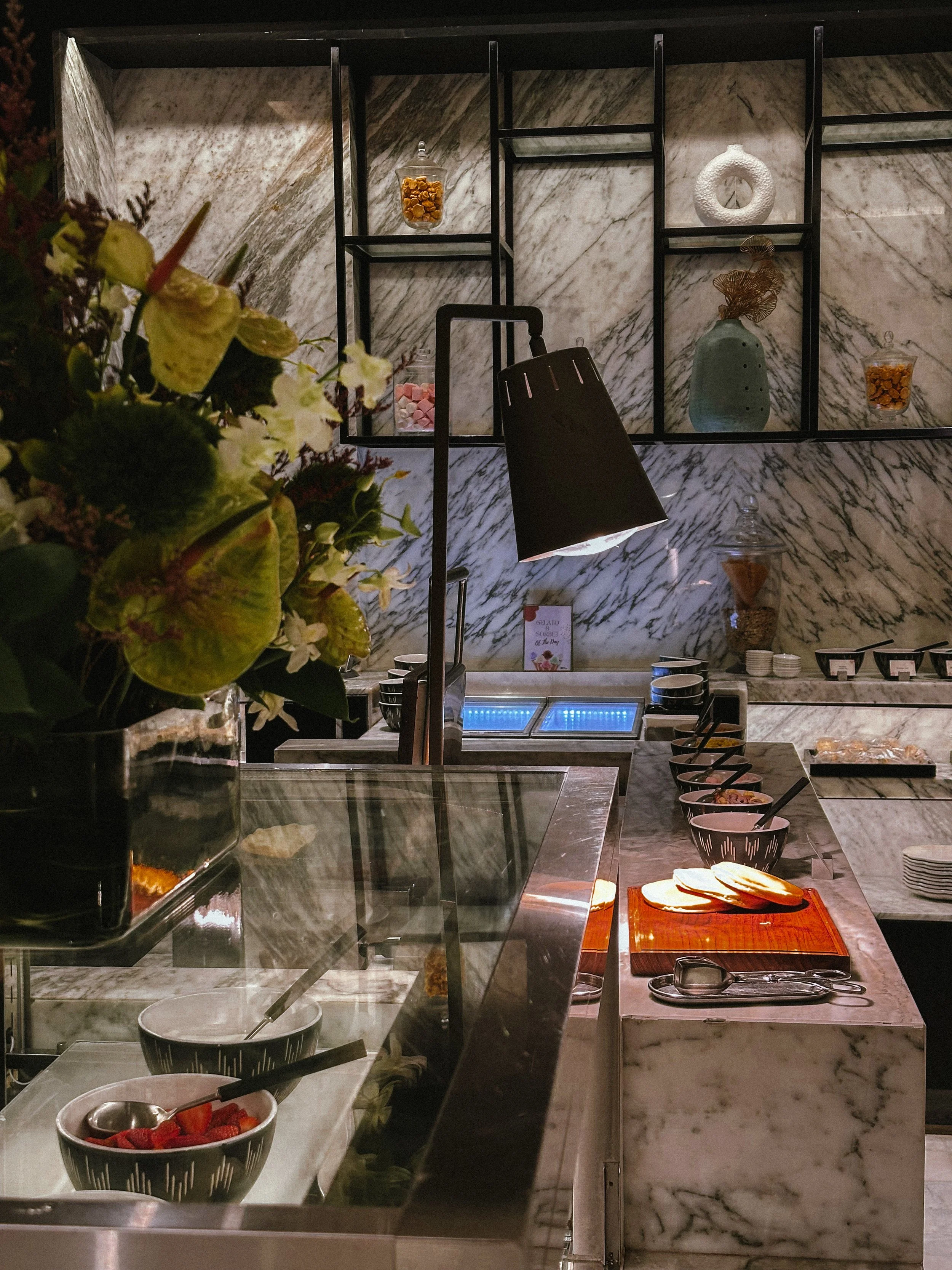 A marble counter with bowls of toppings, a cutting board with sliced bread, and a coffee machine in a modern cafe with decorative shelves and a black lamp.