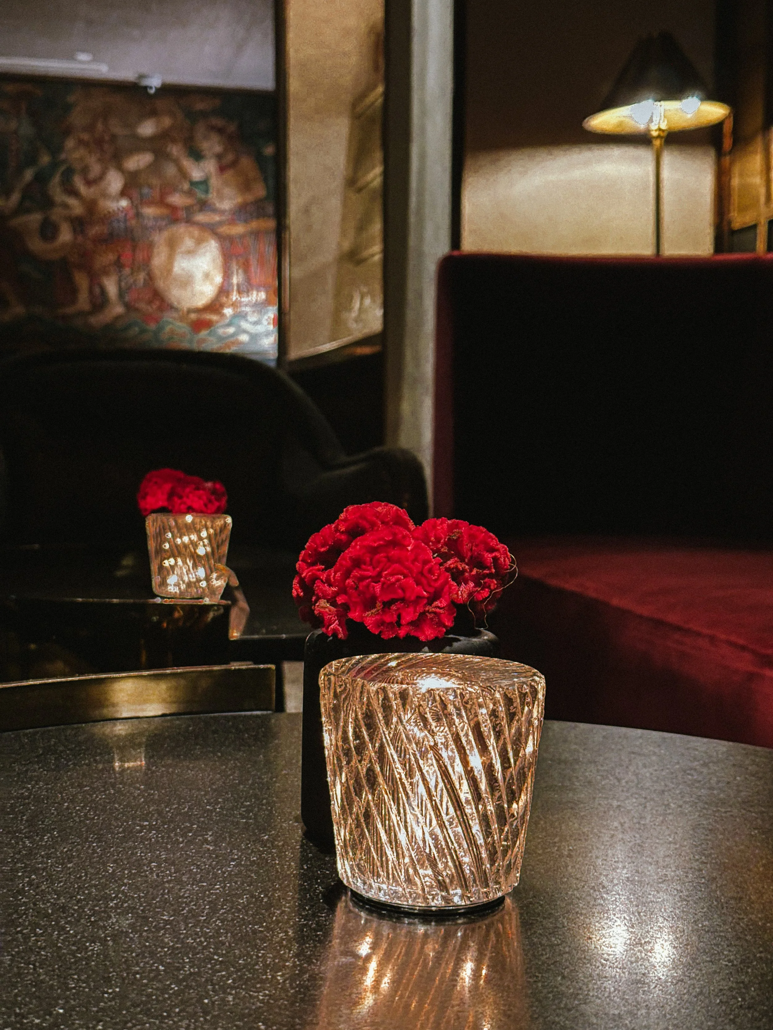 A glass candle holder with a lit candle and a small red flower on a dark table in a dimly lit lounge or restaurant setting.
