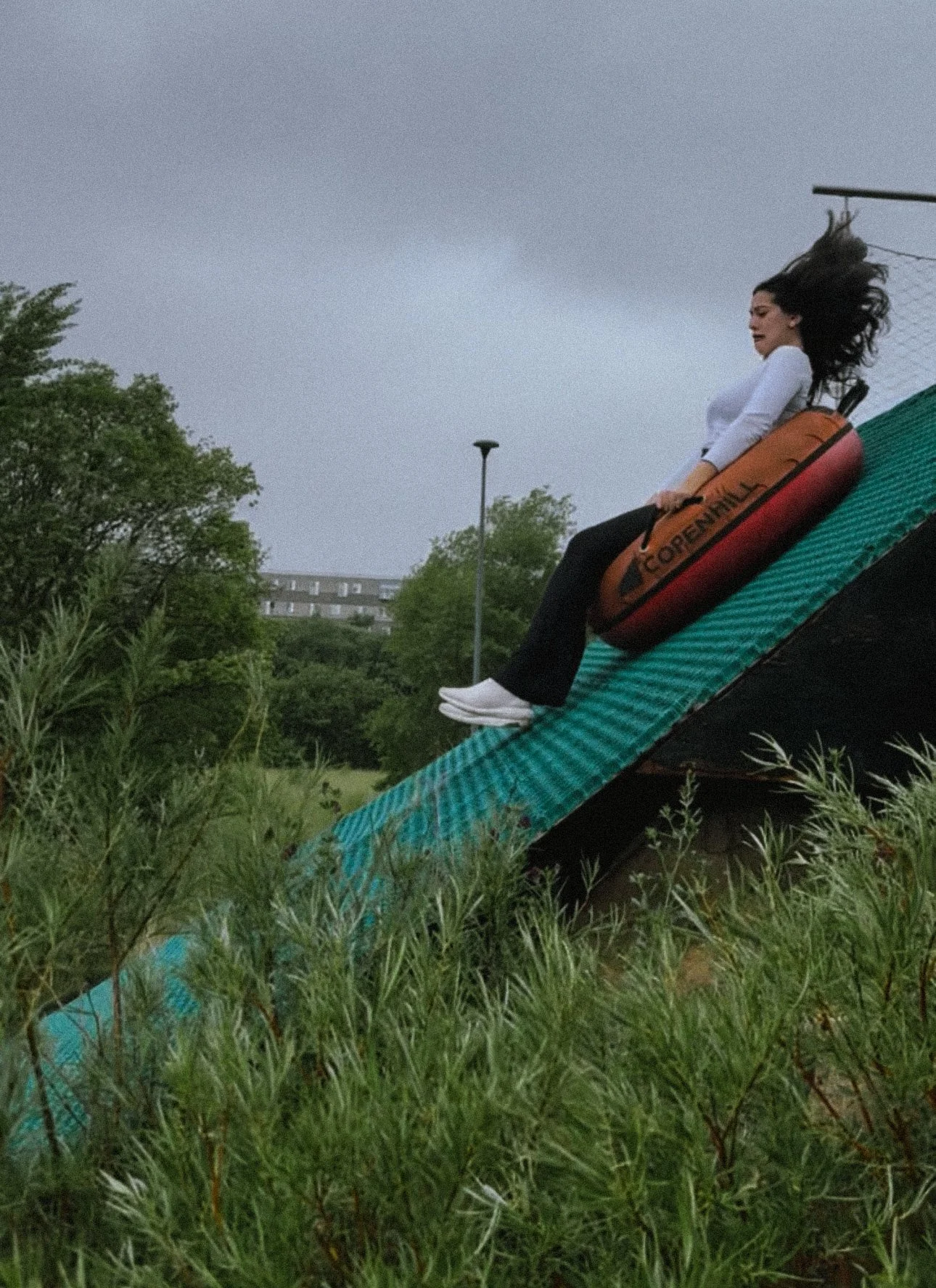 A woman with dark hair sitting on the edge of a green slide, holding a kayak labeled 'Copenhagen,' with a gray sky and trees in the background.