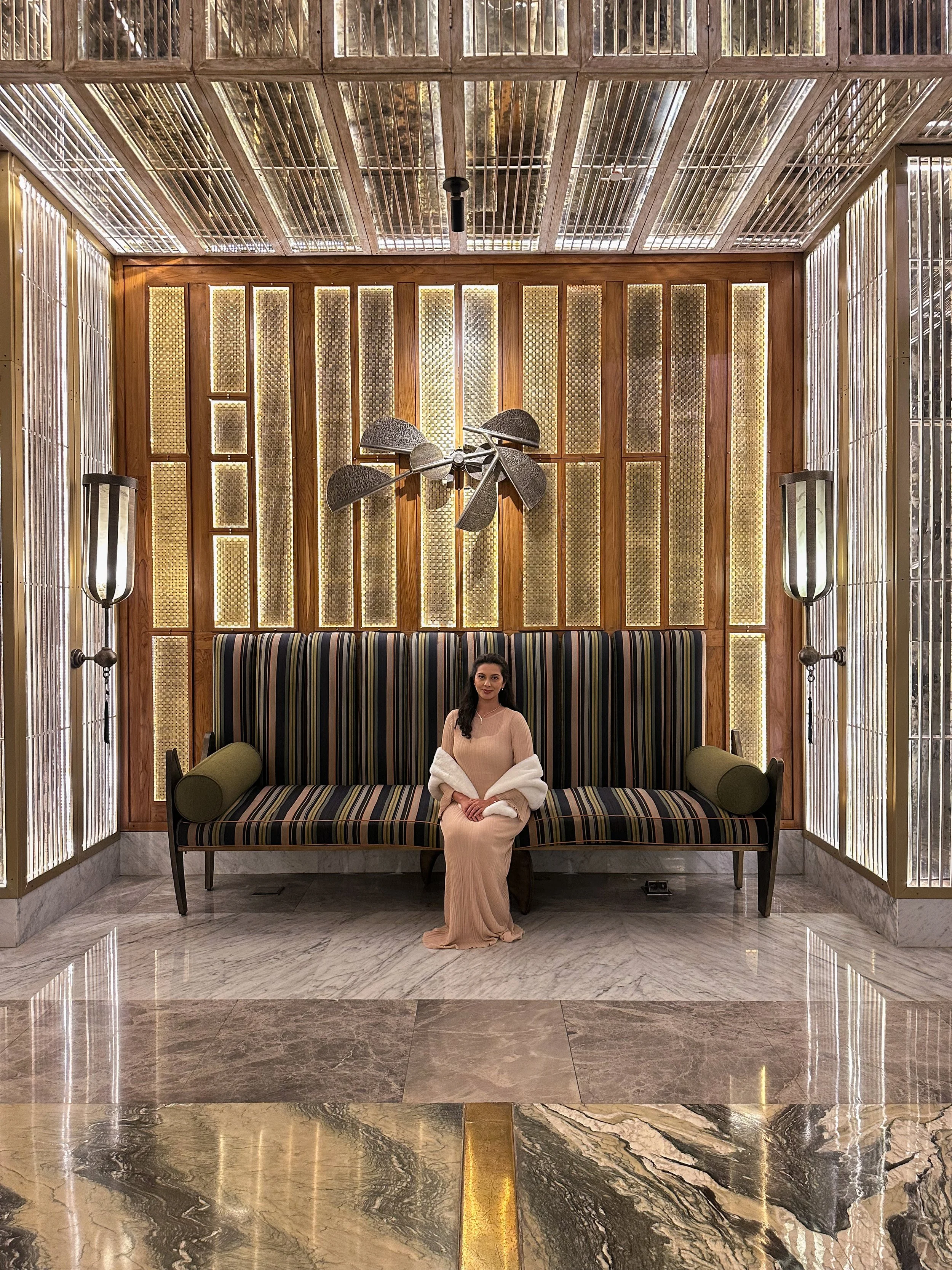 A woman in a tan dress and white shawl sitting on a striped sofa in a luxury interior space with marble floors and decorative lighting